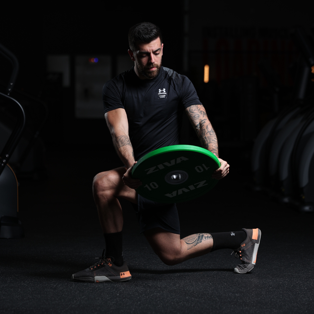 A man with tattoos, wearing black athletic clothing and shoes, kneeling on one knee on a dark gym floor, holding a green weight plate.