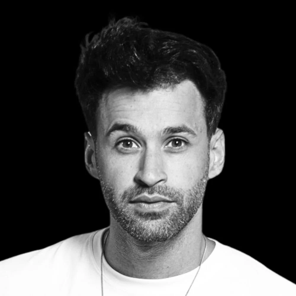 Black and white portrait of a man with dark hair and a beard, wearing a white shirt, against a dark background.