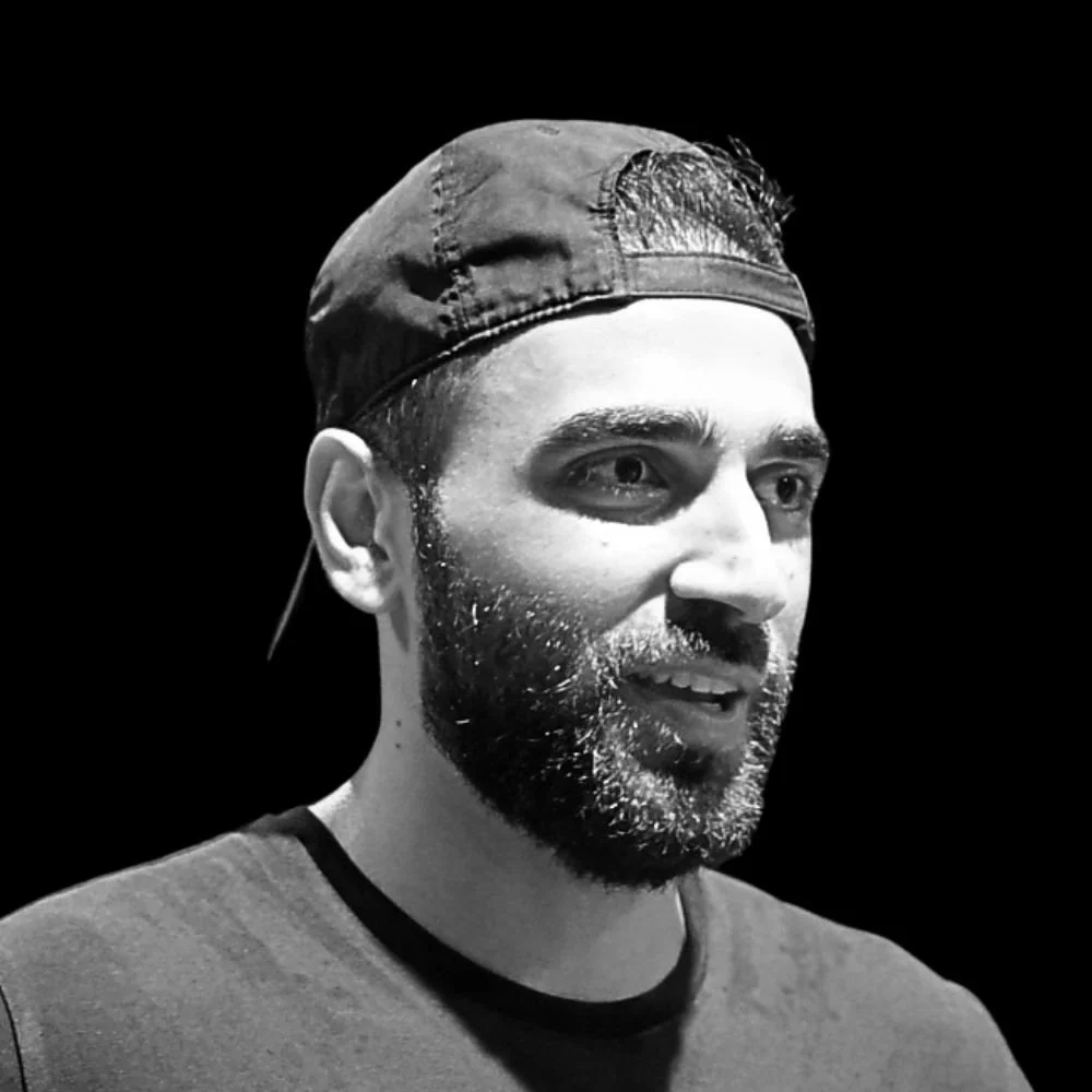 Black and white photo of a man with a beard wearing a baseball cap turned backwards, looking to the right and smiling.