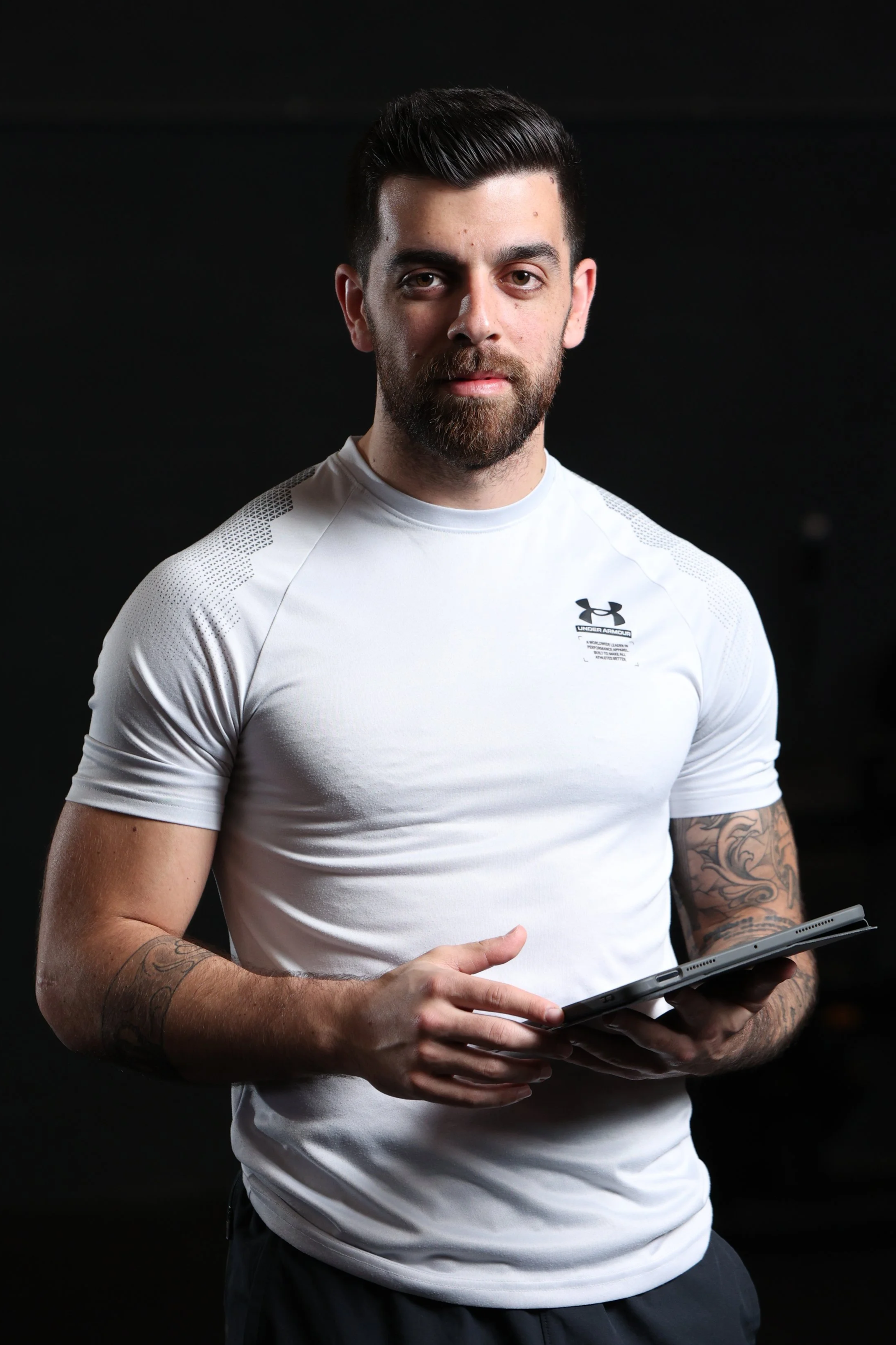 A man with dark hair and a beard wearing a white Under Armour t-shirt, holding a tablet in his hands, standing against a dark background.