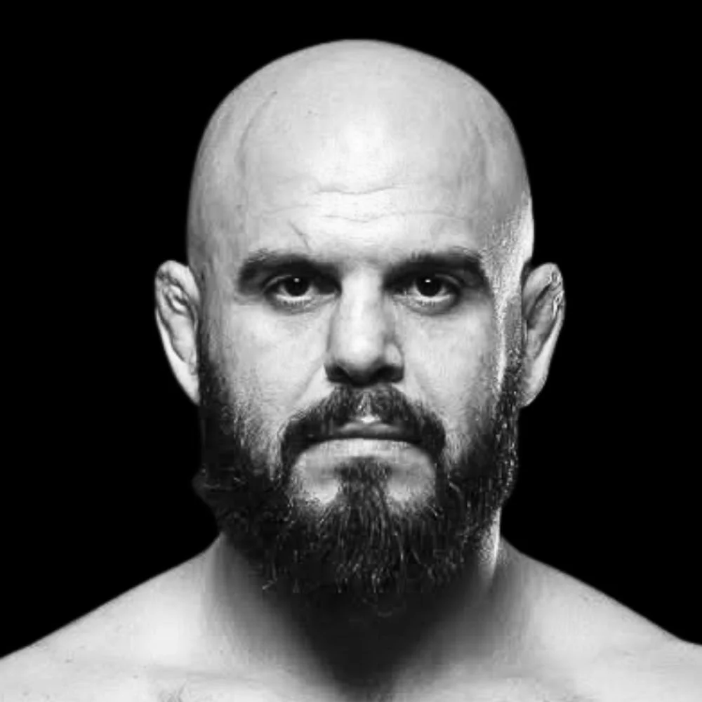 Black and white portrait of a man with a shaved head, a full beard, and a serious expression, shirtless with a black background.