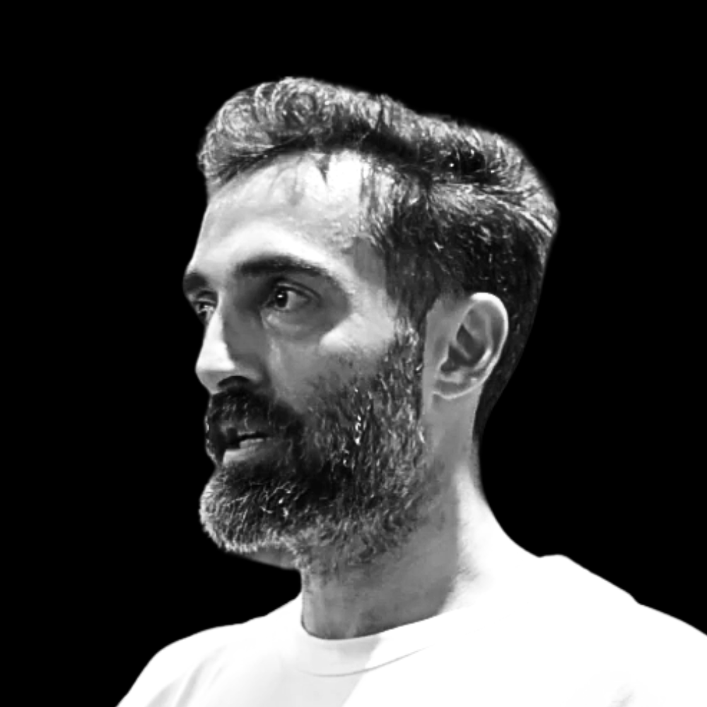 Black and white portrait of a man with a beard and styled hair, looking to the side against a dark background.