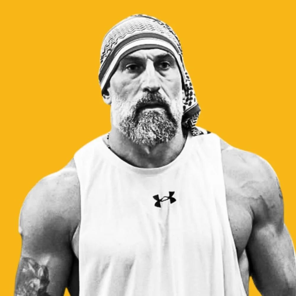 A black and white photo of a muscular man with a beard, wearing a headscarf and a sleeveless Under Armour shirt, set against a yellow background.