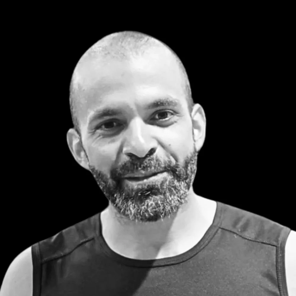 A black and white photo of a man with a beard and short hair, wearing a sleeveless athletic shirt, smiling slightly against a dark background.