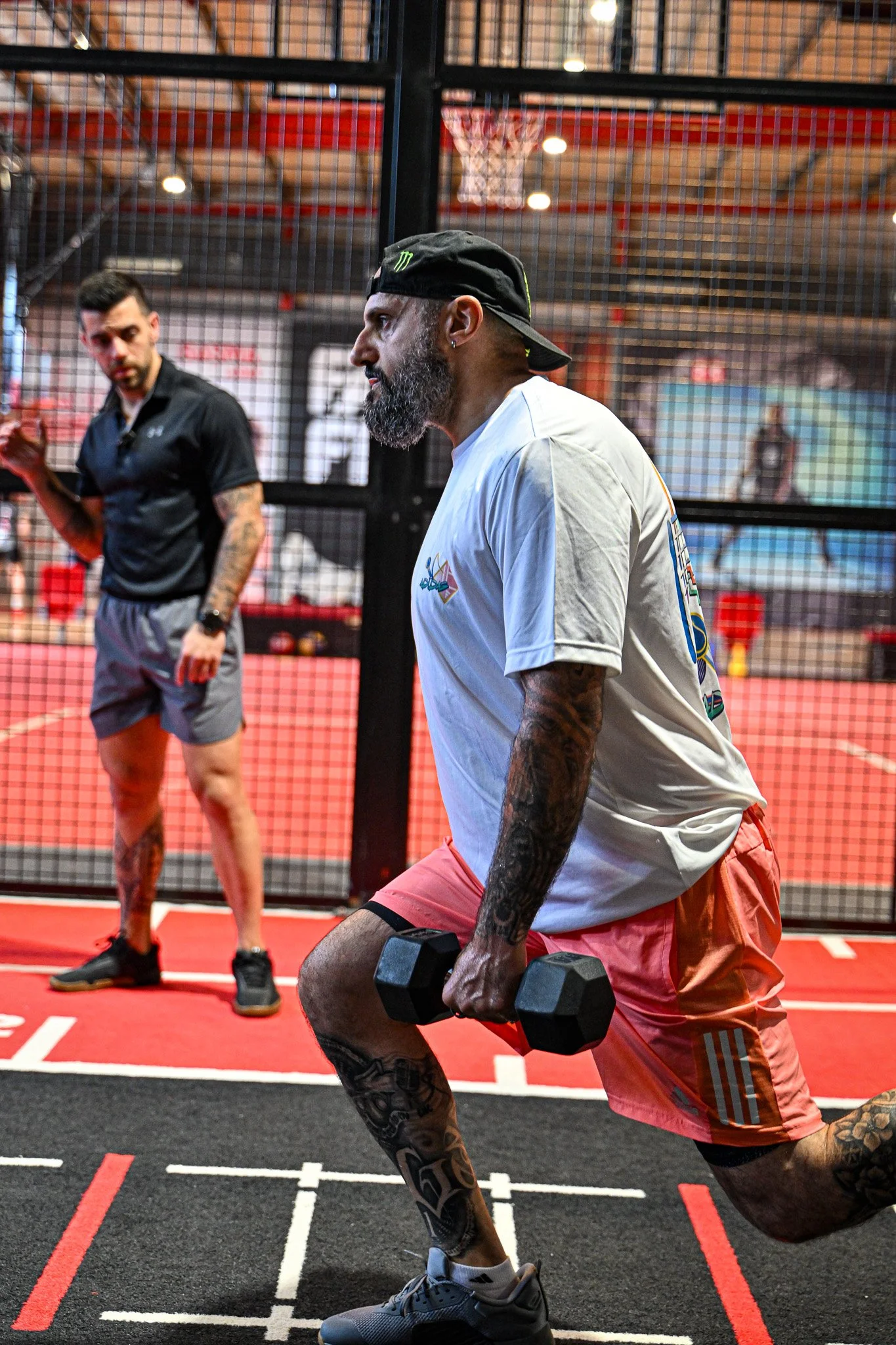 A man with tattoos, beard, and a black cap doing a lunge exercise with a dumbbell in each hand inside a gym, with another man observing in the background.