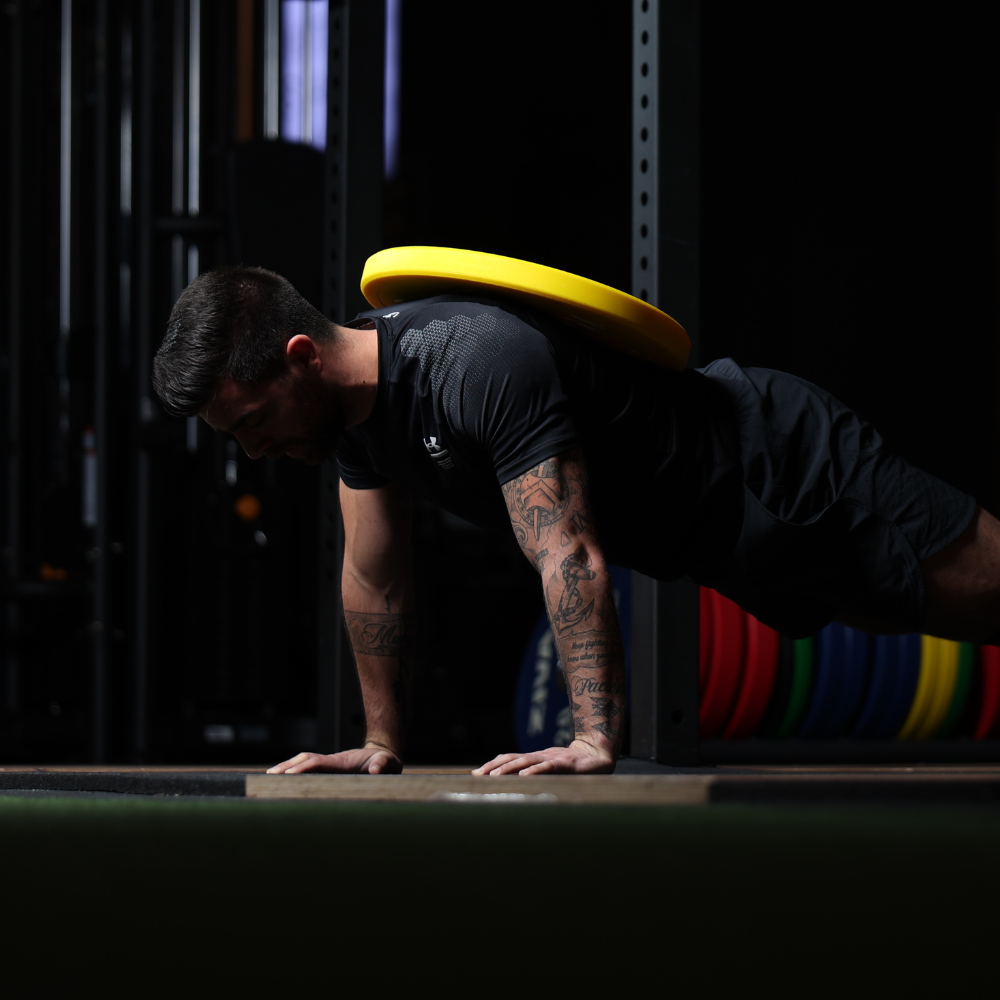 Man doing a push-up with a yellow weight plate on his back in a gym.