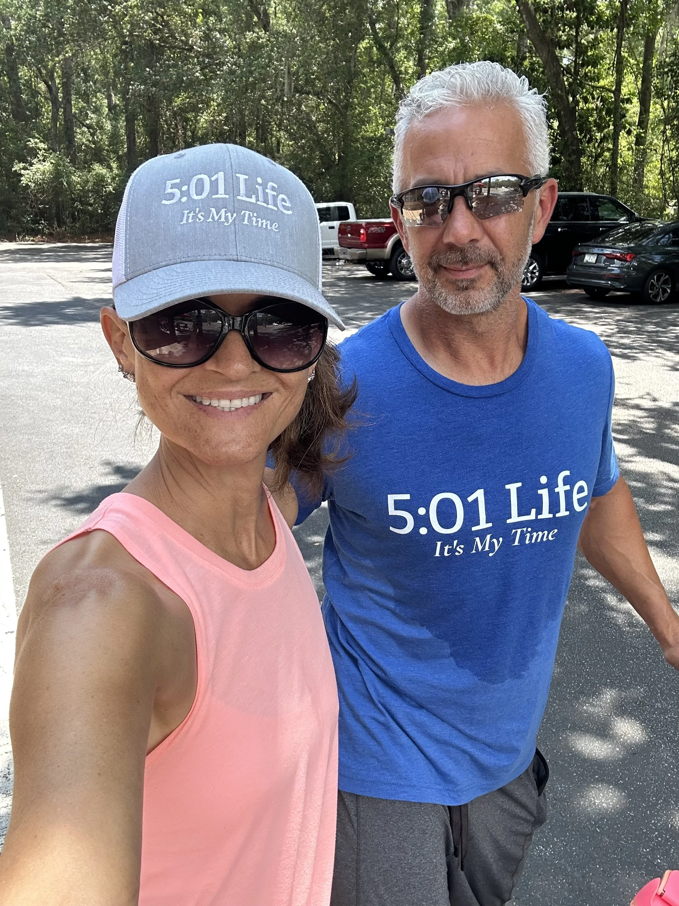 A woman and a man are taking a selfie outdoors in a parking lot surrounded by trees. Both are wearing sunglasses and matching shirts with the text '5:01 Life It's My Time.' The woman is wearing a pink sleeveless top and a light blue baseball cap, while the man is dressed in a blue t-shirt and gray shorts.
