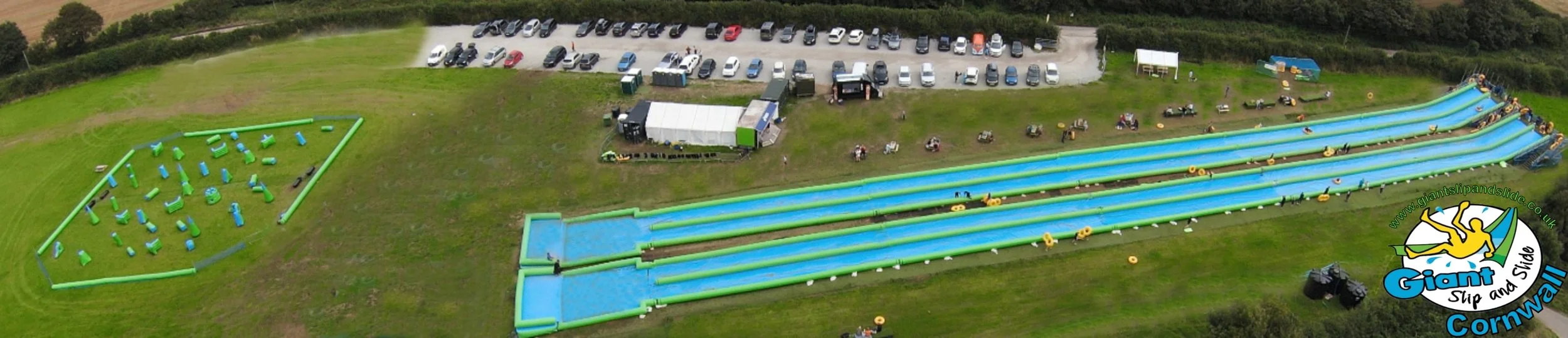 Aerial view of the large outdoor slip and slide park featuring four of the 7  water slides, a separate gravel area with parked cars,  and a water play area with inflatable obstacles. The park logo reads 'Giant Slip and Slide Cornwall'.