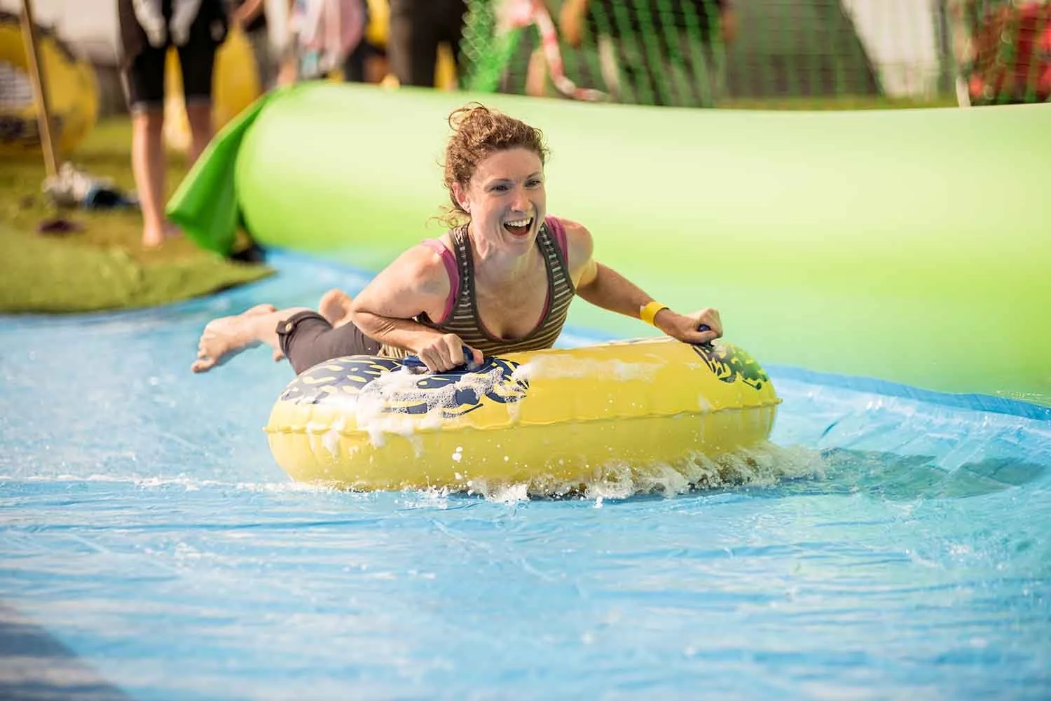 Woman riding a yellow inflatable down the slip and slide smiling and enjoying a water slide ride at an amusement park.