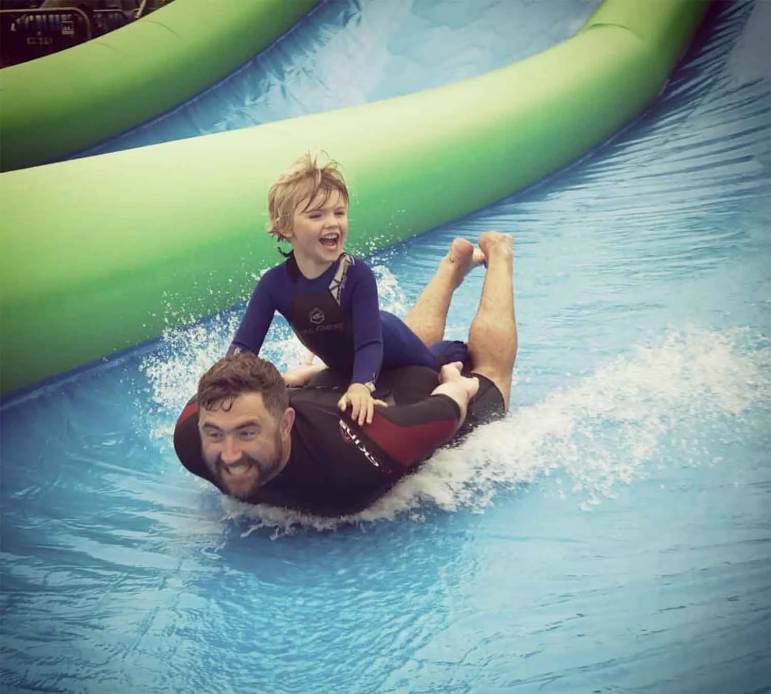 A young boy lying on his stomach on an adult man's back, riding a water slide with blue water and a green inflatable barrier, both smiling and having fun.