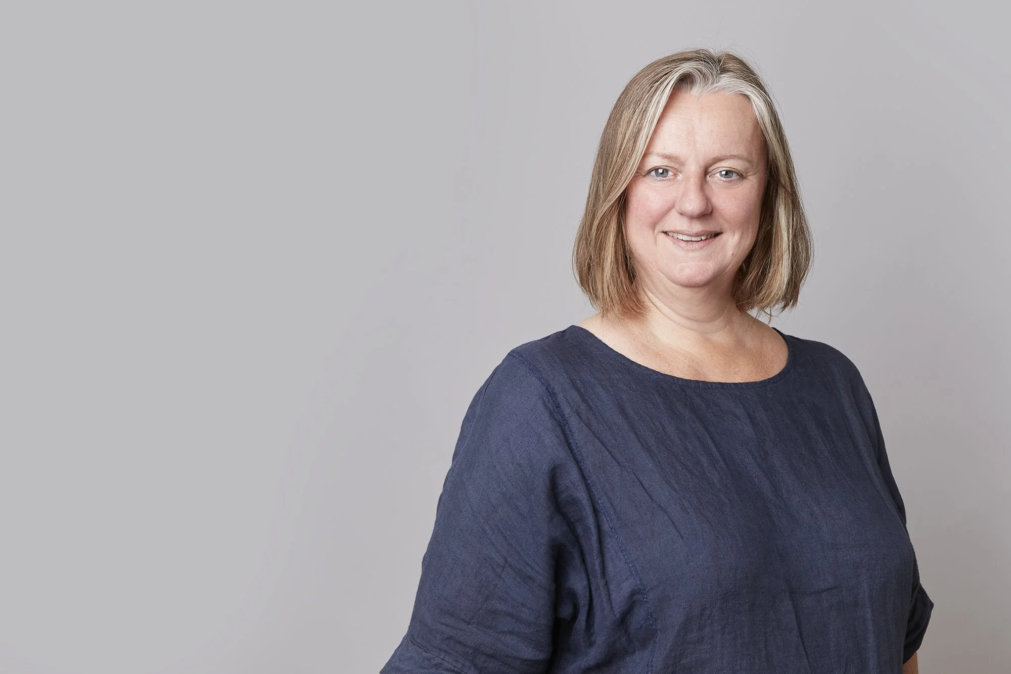 A woman with shoulder-length gray hair, blue eyes, and earrings, wearing a dark blue top, standing in front of a brick wall.