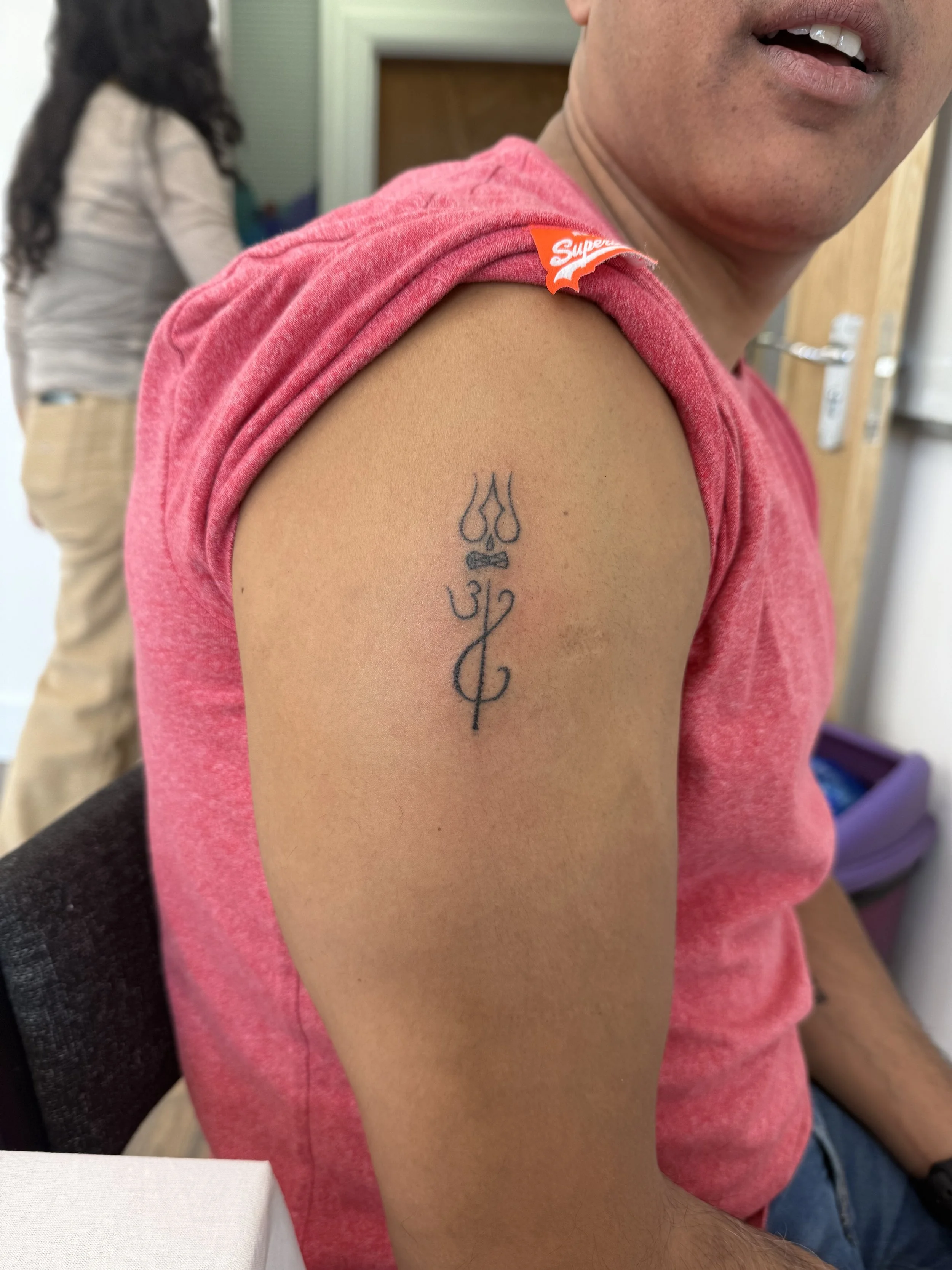 A man with a tattoo on his upper left arm, showing a design with three lines, a trident, a bow, and a stylized number '2', wearing a pink T-shirt with a 'Supreme' tag on the sleeve. In the background, a woman with curly hair in a beige long-sleeve shirt and khaki pants, and a short blurred view of a person, a door, and a stored container.