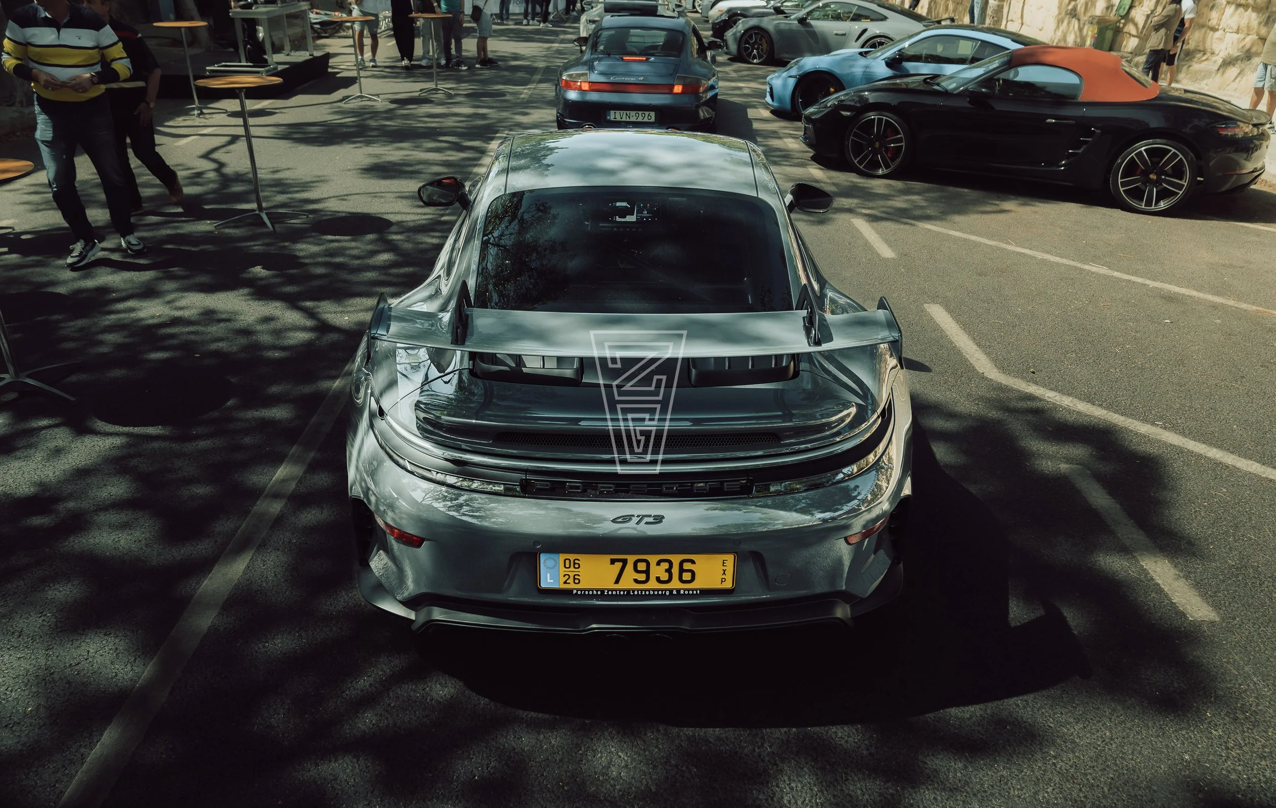 A silver Porsche 911 GT3 parked in a lot with other sports cars, people walking around and trees casting shadows.