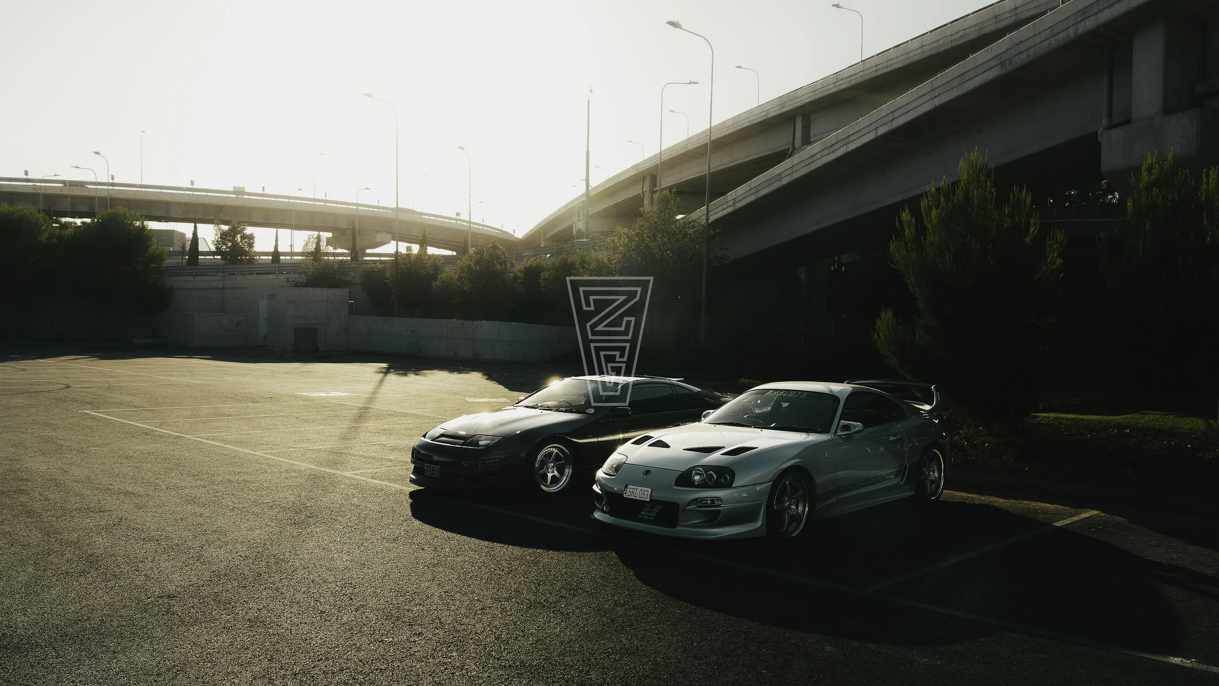 Two sports cars, a black one and a silver one, parked in an empty parking lot beneath large overpasses with trees lining the area, during late afternoon sunlight.