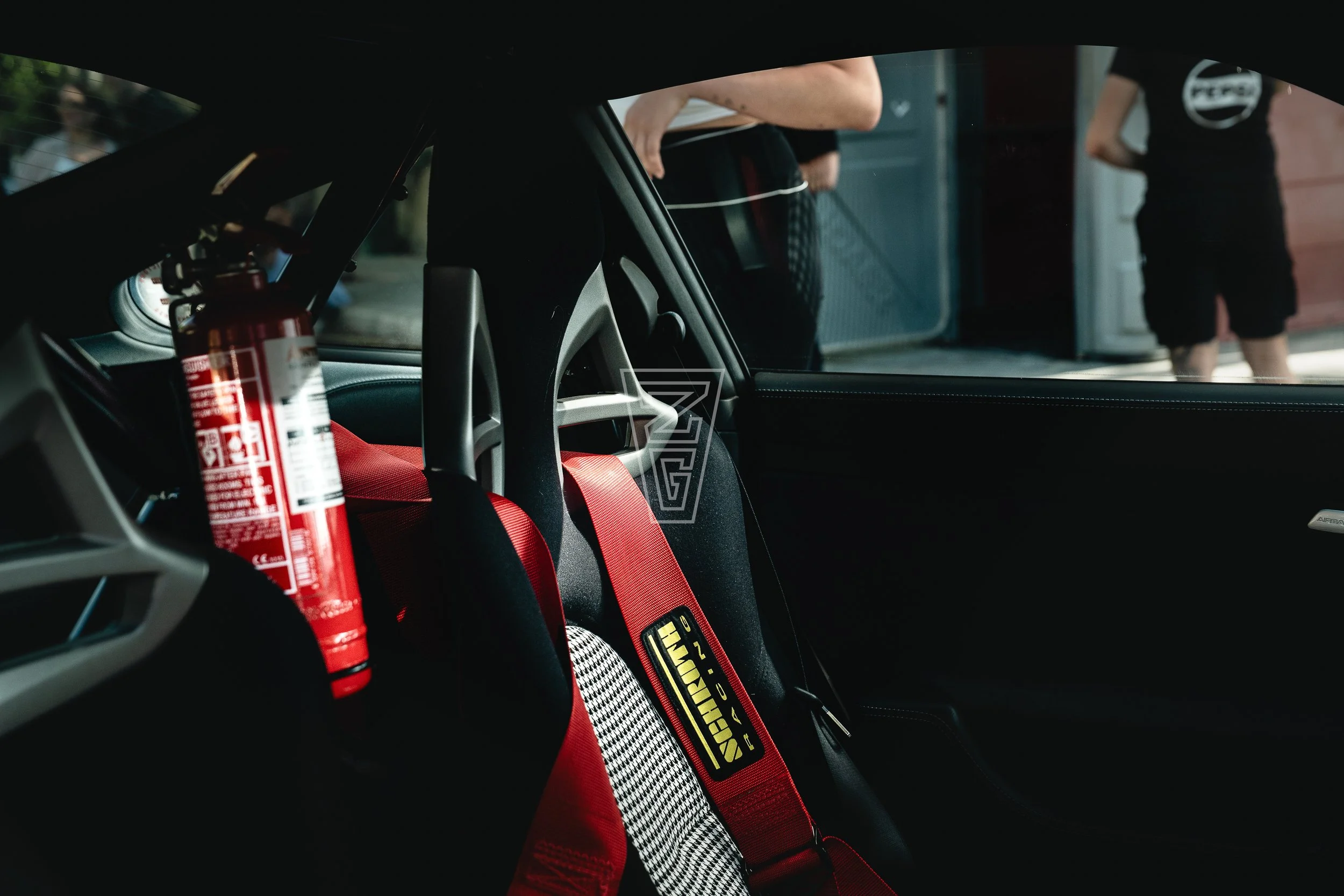 The interior of a car showing a firefighting fire extinguisher mounted next to a racing harness seatbelt with a red and black checkered pattern and the brand 'Schroth' visible.