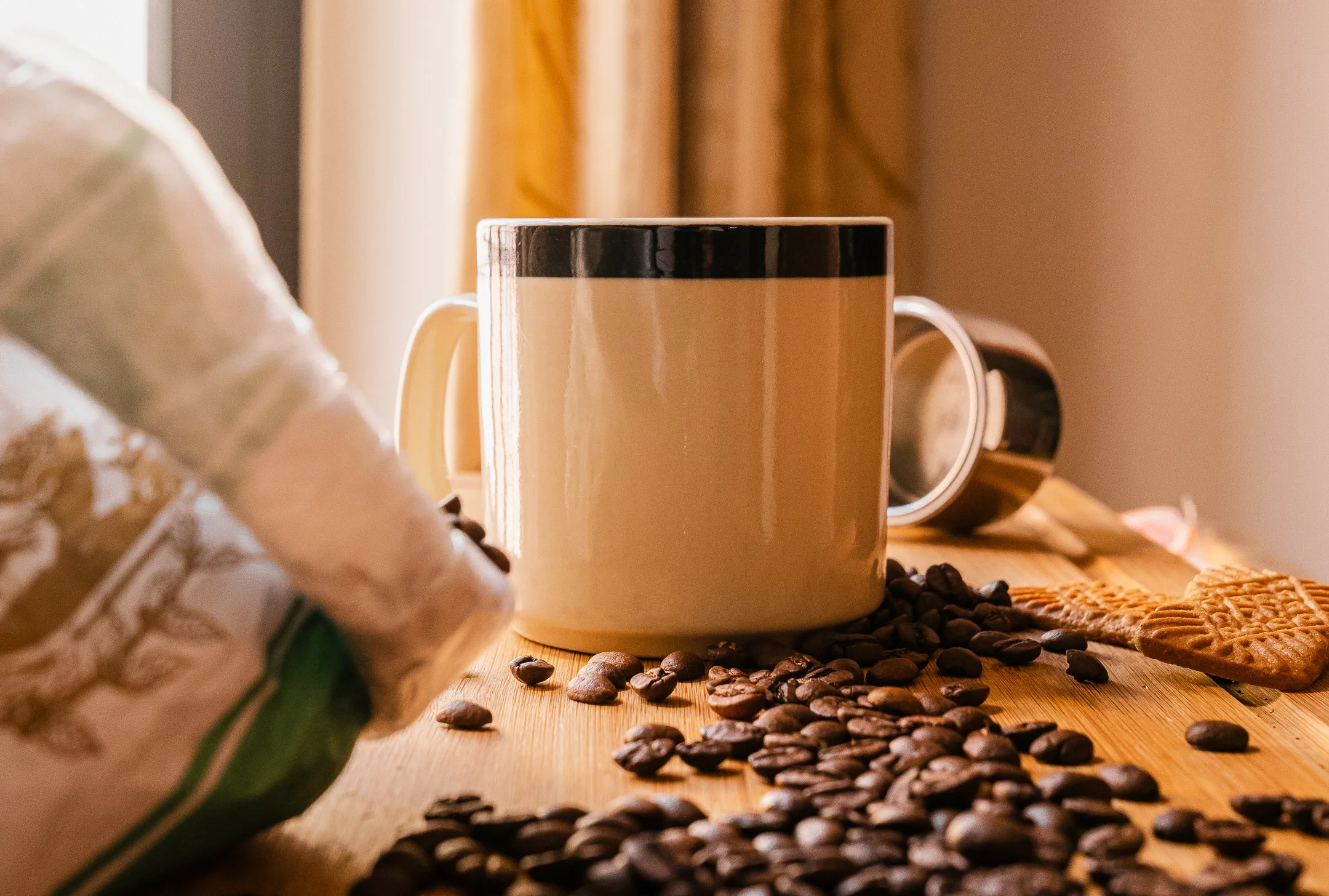 Coffee beans spilled on a wooden table, with a beige mug, a bag of coffee, a stainless steel canister, cookies, and a yellow cloth in the background.