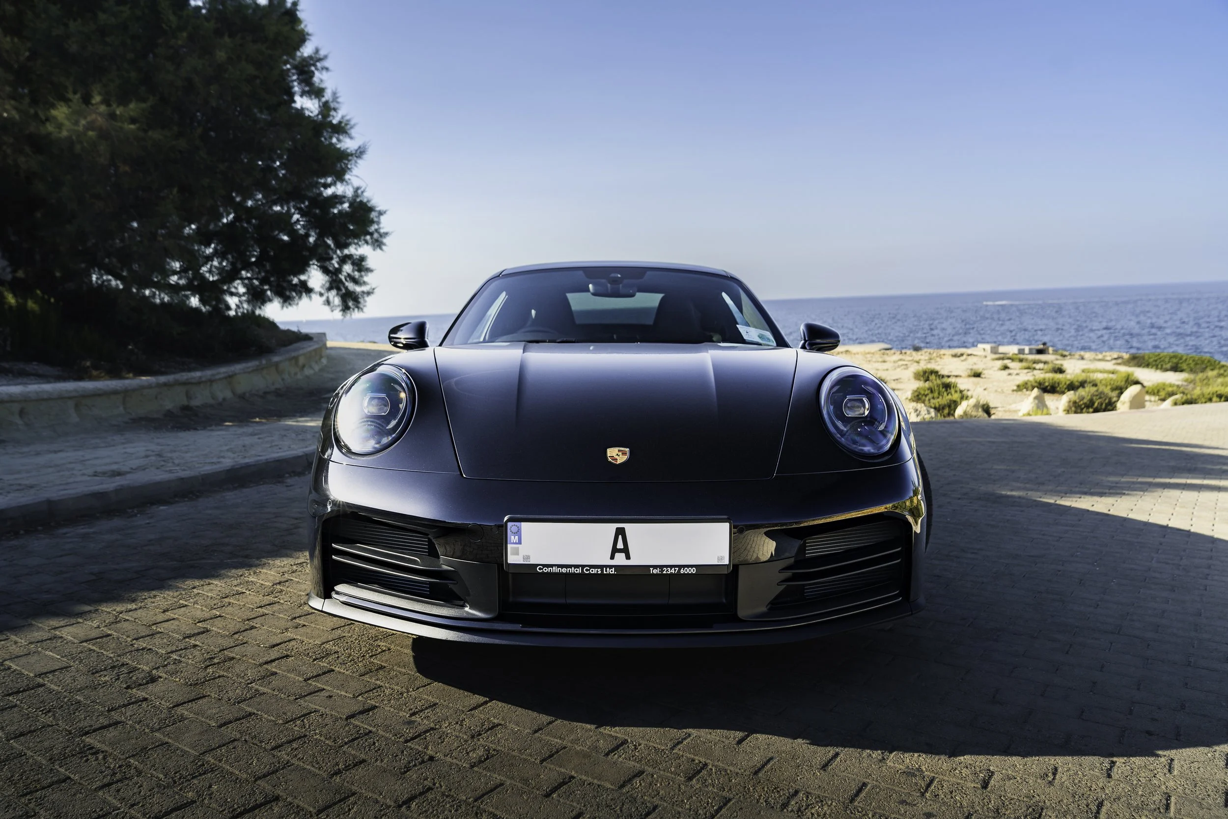 Front view of a black Porsche sports car parked on a paved area near the coast, with trees on the left and the sea in the background under a clear blue sky.