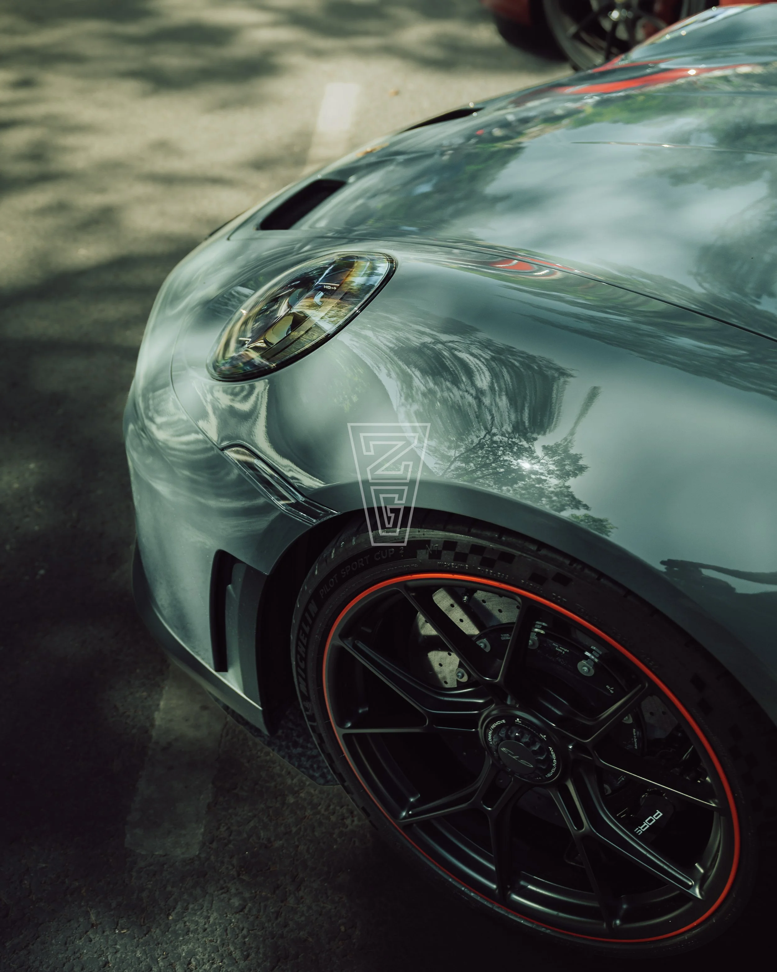 Close-up of a silver sports car with black wheels and a red trim, showing the front headlight and part of the hood and wheel, parked on asphalt.