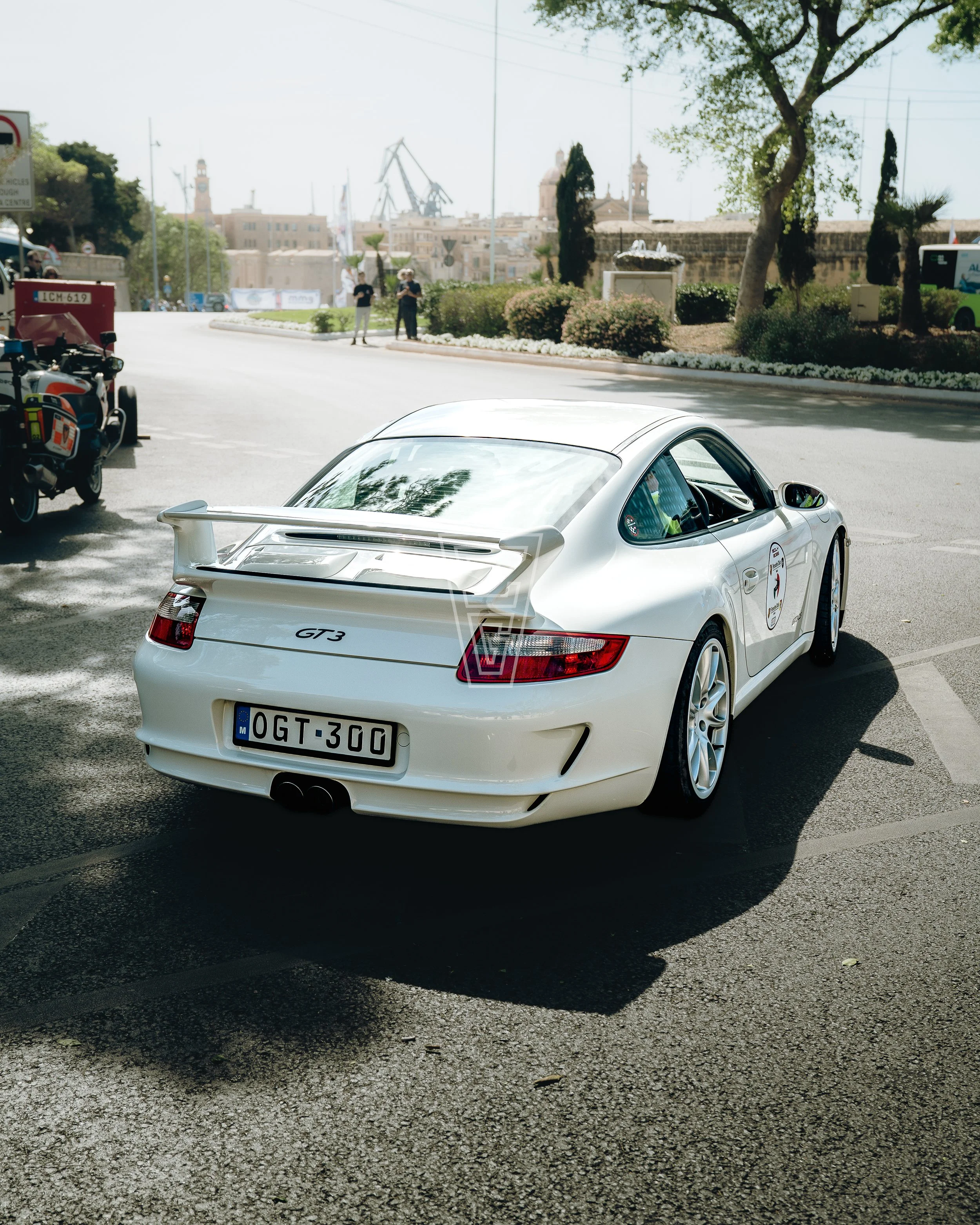 A white Porsche 911 GT3 sports car parked on a street with trees, bushes, and a fountain in the background. Other vehicles and people are visible in the background.