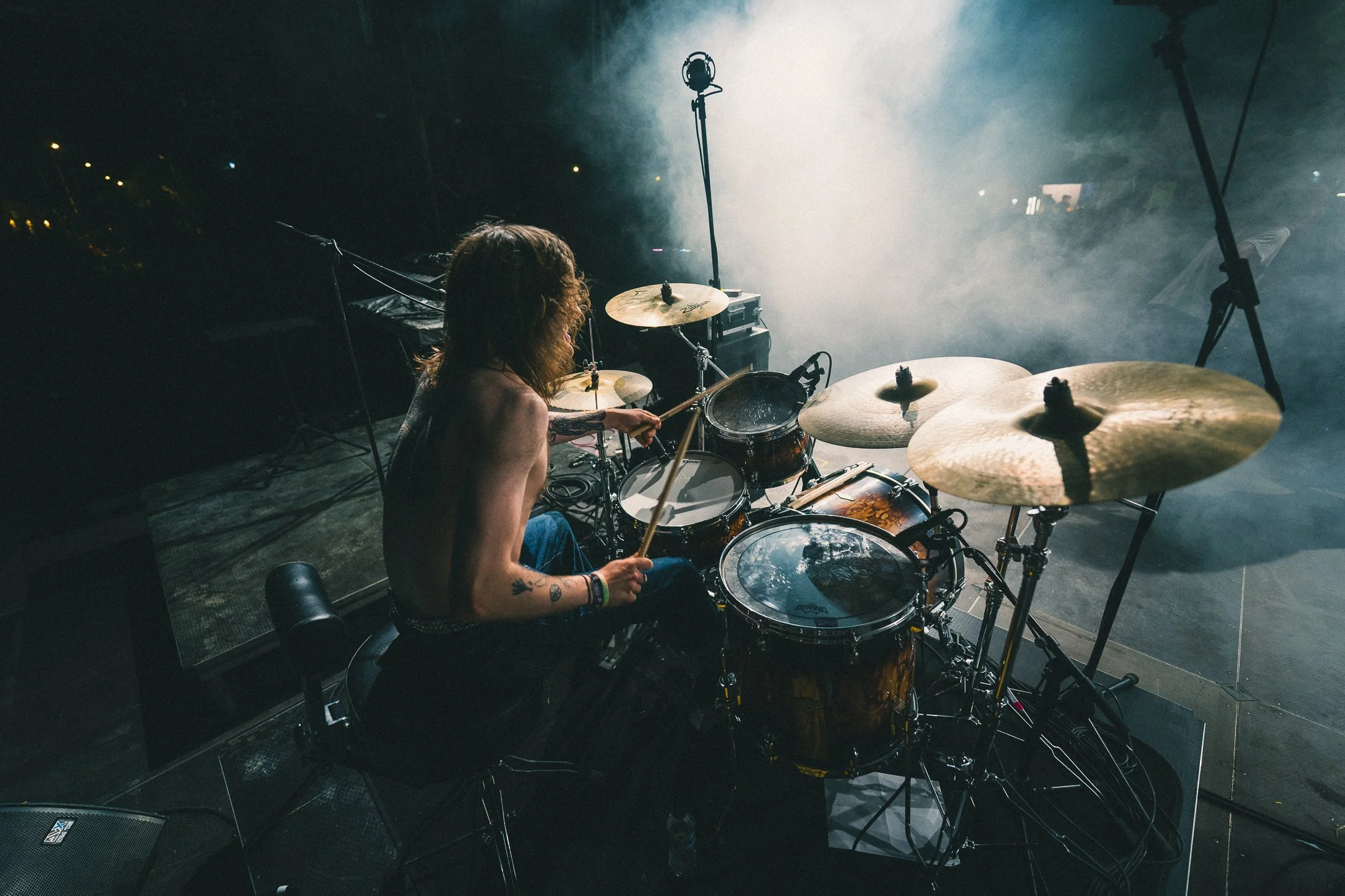 Shirtless drummer performing on stage surrounded by theatrical smoke and intense spotlights.
