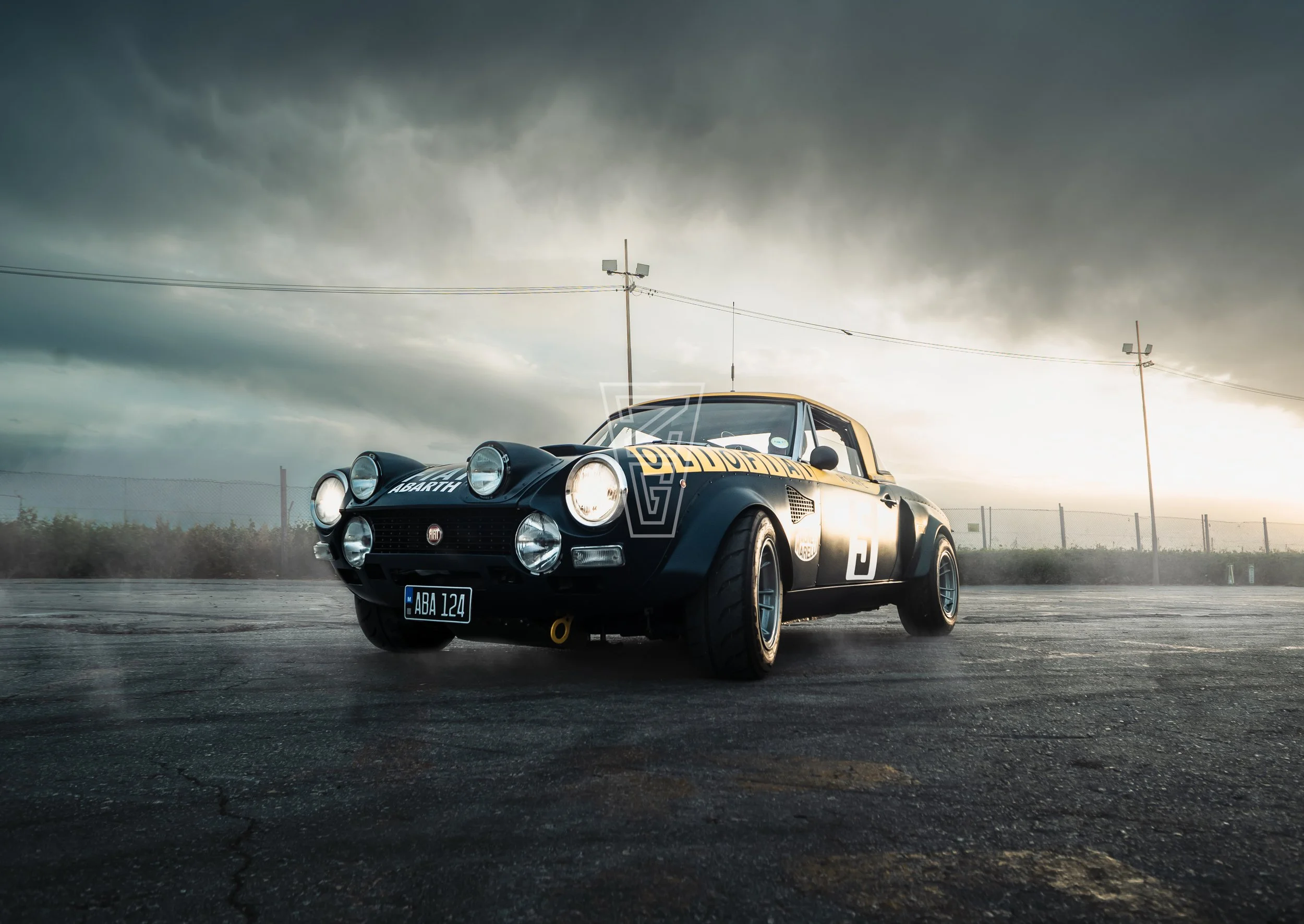 A vintage black sports car with racing decals, including the number 37, is parked on an outdoor lot under a dark, cloudy sky. The car features a Fiat badge and multiple headlights, with a license plate reading 'ABA 124.'