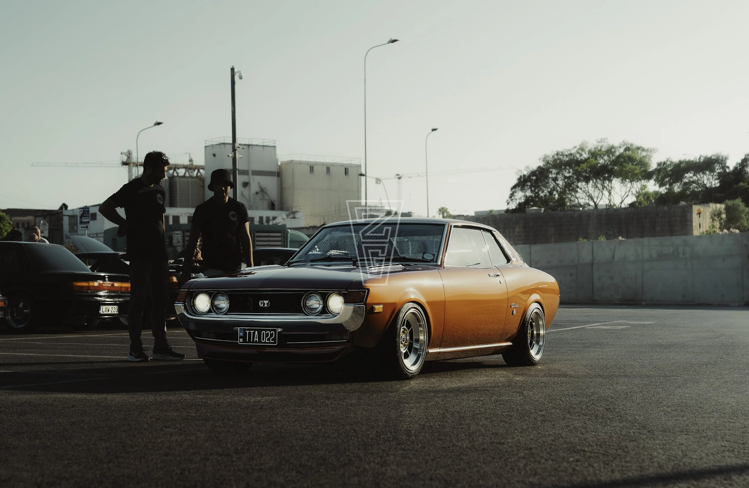 A classic orange muscle car parked in a lot with two men standing nearby, one wearing a bucket hat, at sunset with trees and buildings in the background.