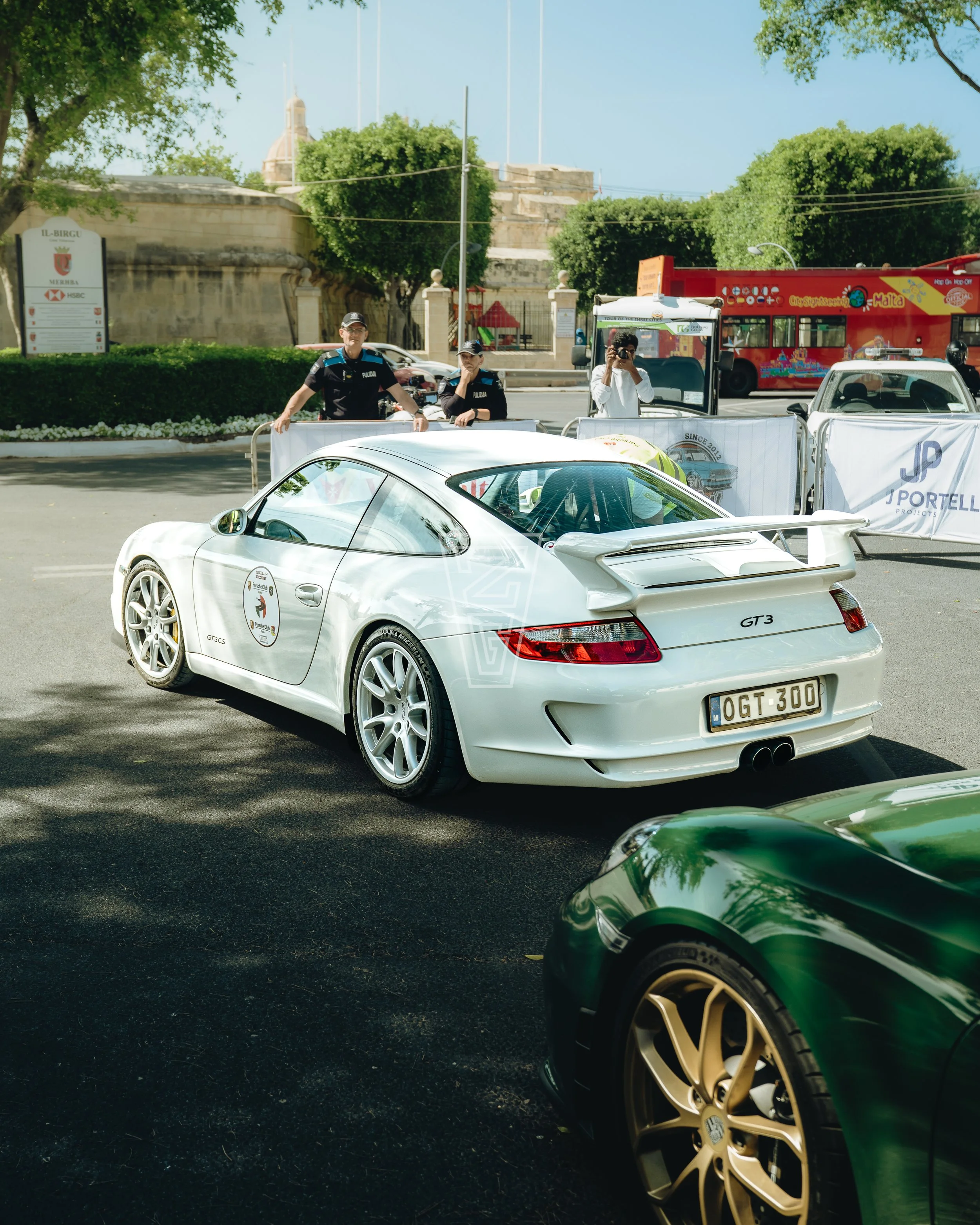 White sports car with Porsche logo, parked on street with two police officers and another person taking a photo in front of a historic building and a red tourist bus, in a sunny city scene.
