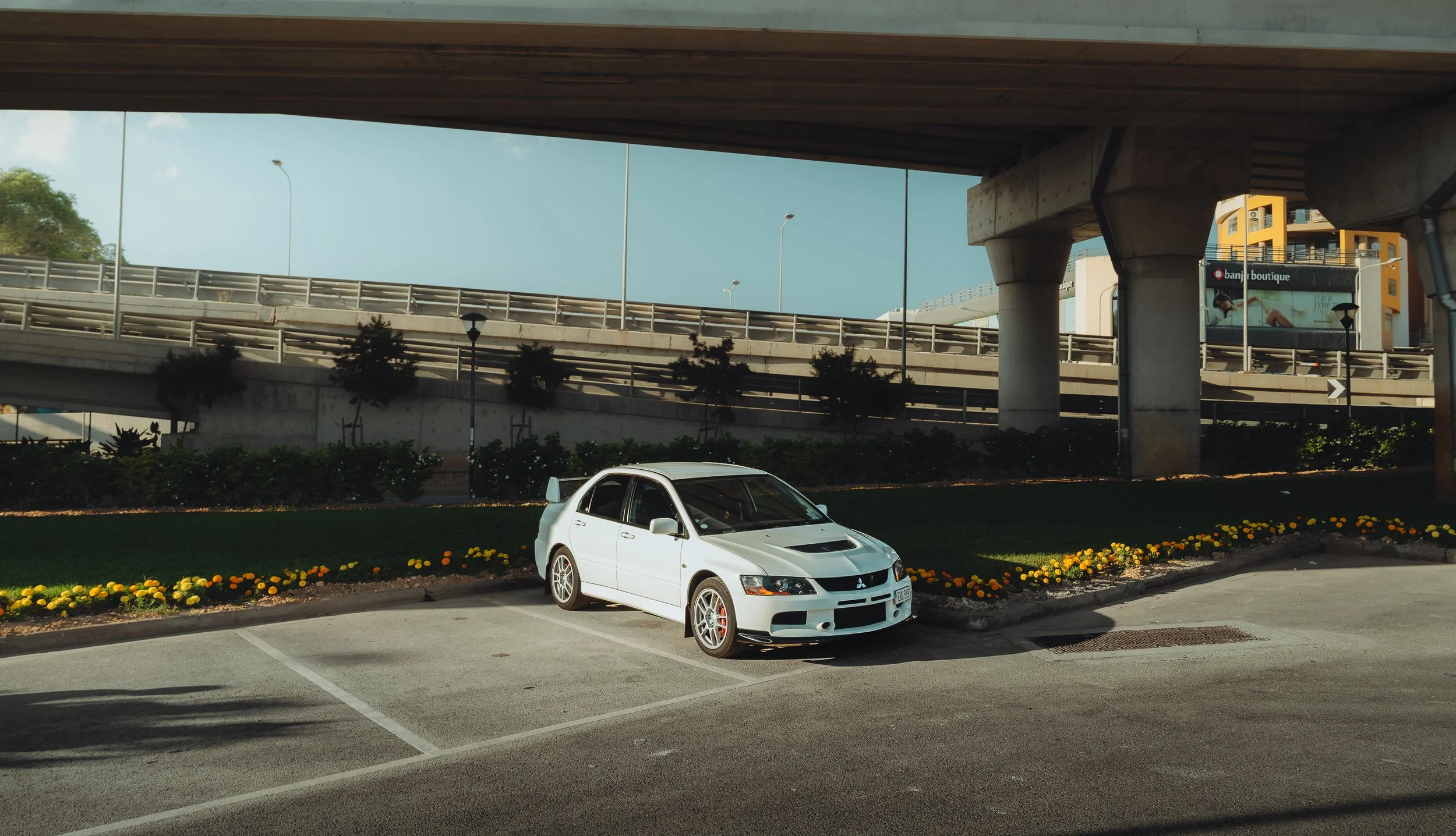 A white Mitsubishi Lancer Evolution car parked in a designated parking space in front of a landscaped area with yellow flowers, under a large overhead highway or bridge structure with multiple support columns. There are trees, lampposts, and a billbo