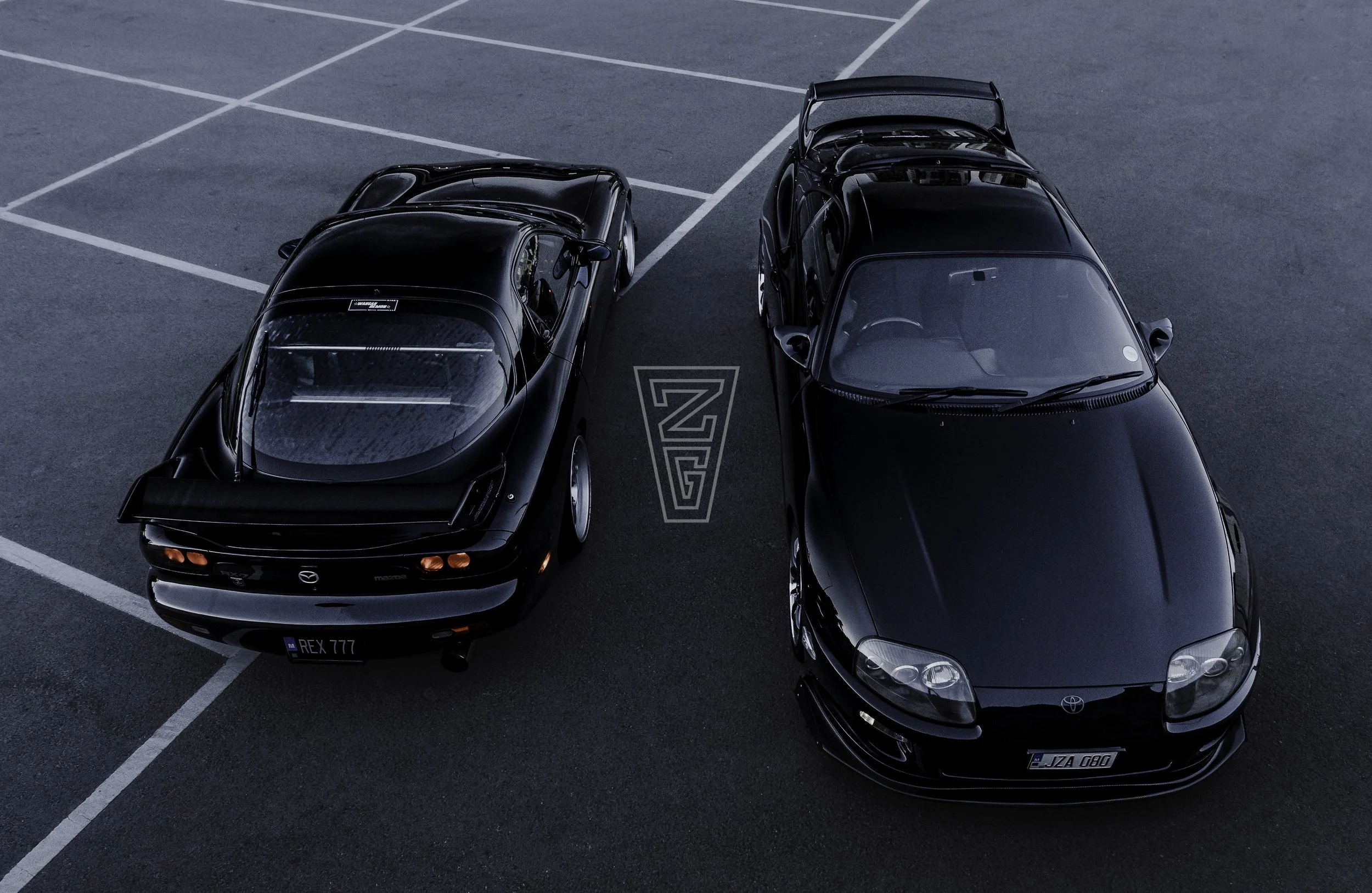 Two black sports cars parked side by side in an empty parking lot with white painted lines and a dark asphalt surface.