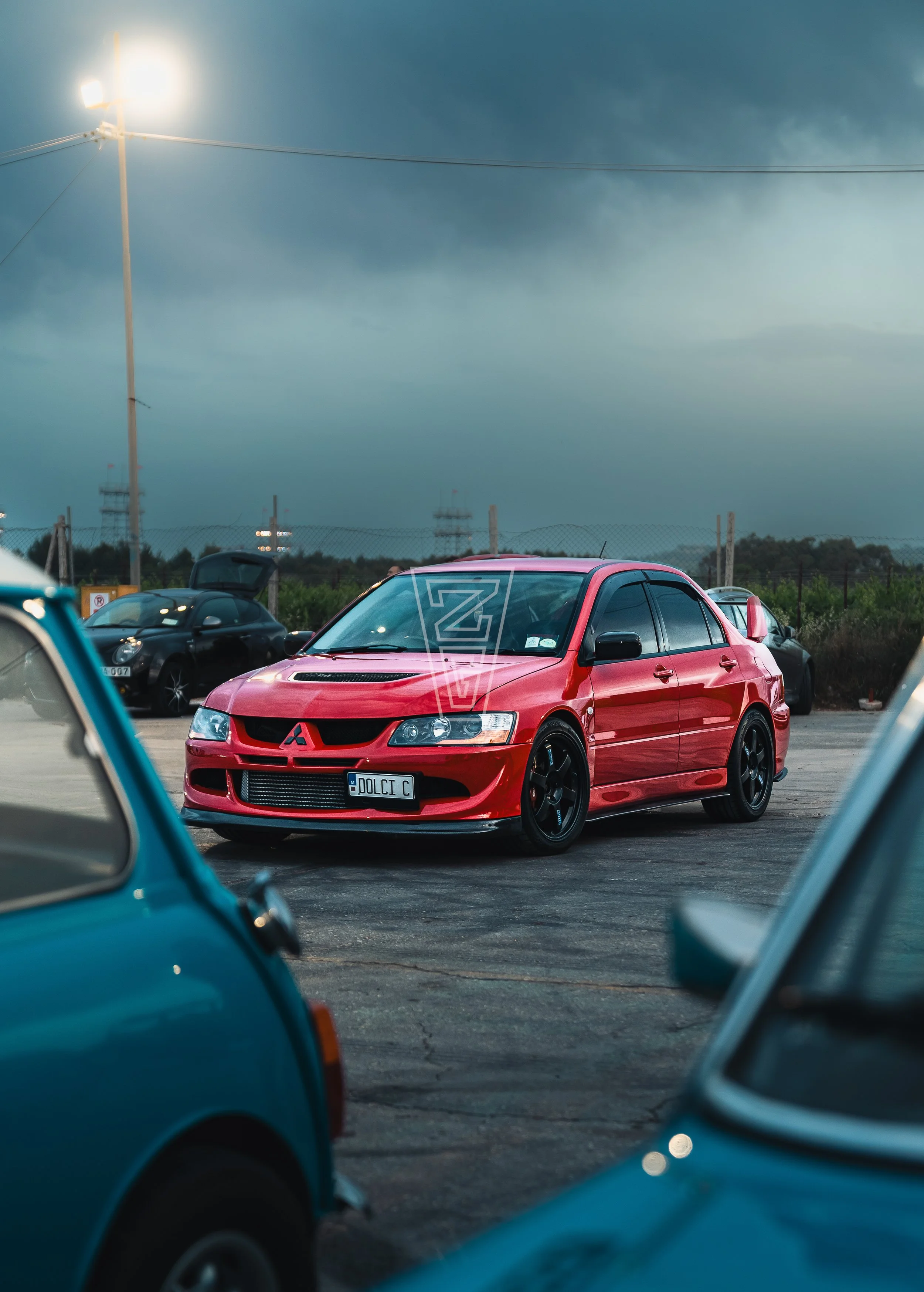 A red Mitsubishi Lancer with a custom license plate reading 'DOLCI C' parked among other cars in a parking lot under dark stormy clouds, with a tall streetlight illuminating the scene.