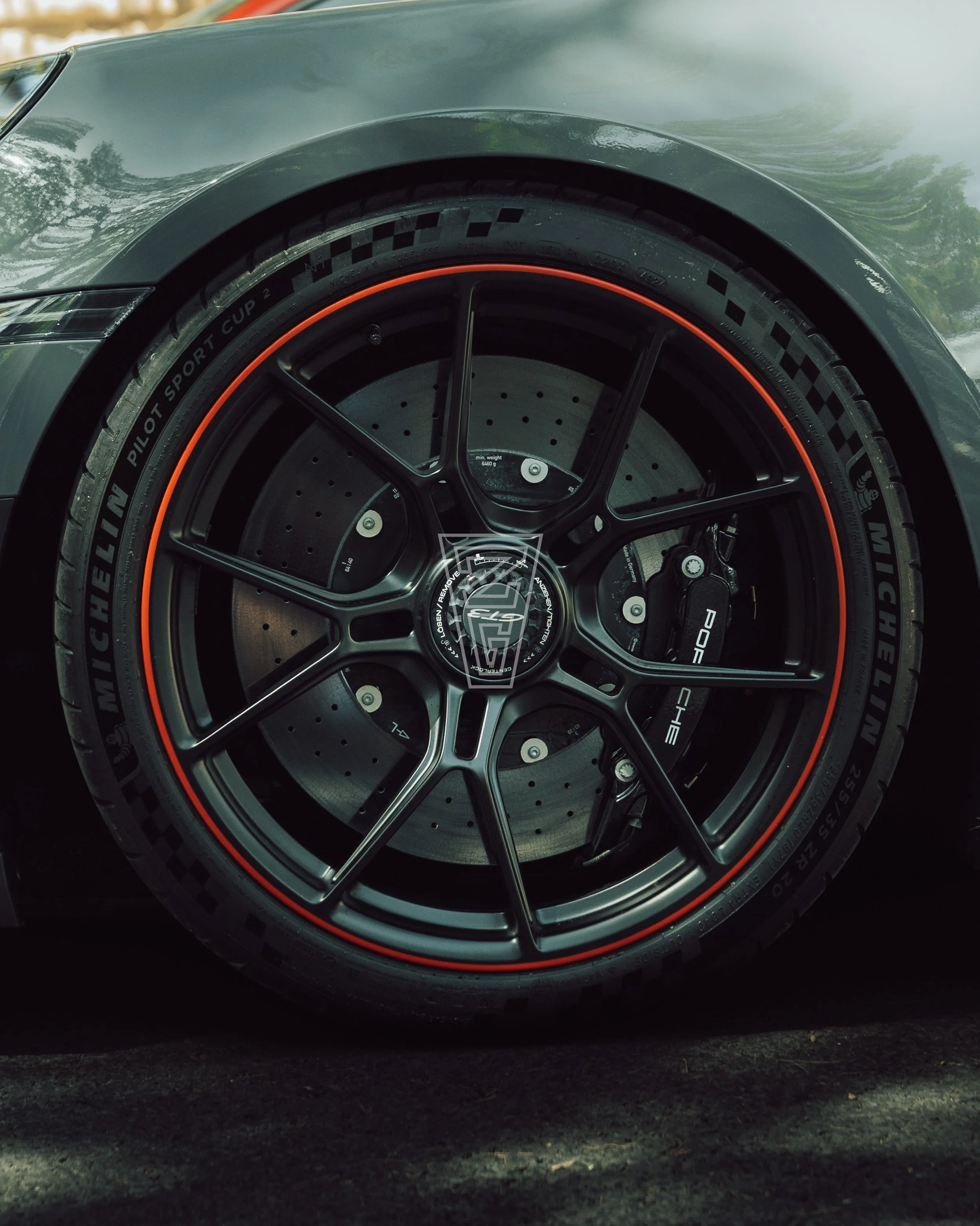 Close-up of a car tire with black wheel rim, orange accent, and brake disc, on a black asphalt surface.