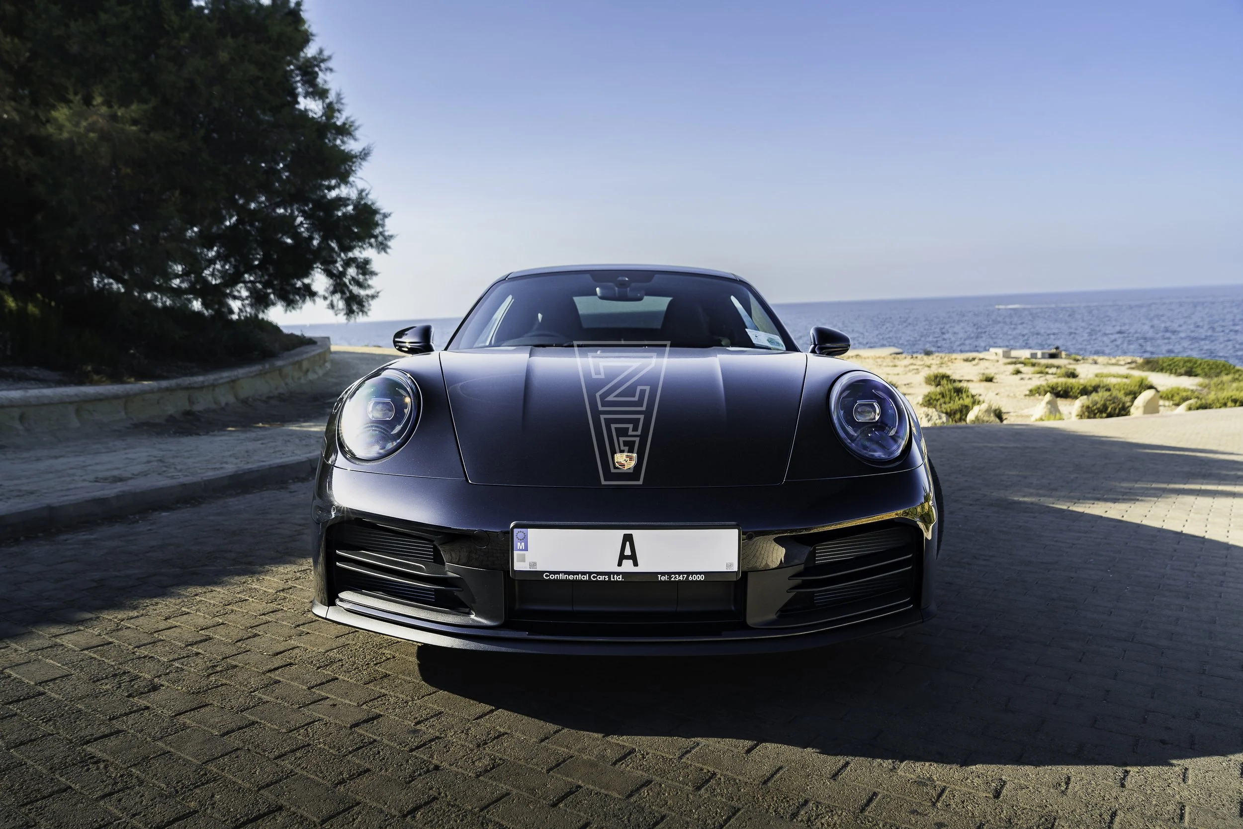 Black Porsche sports car parked near the seaside with the ocean in the background, leaves of a large tree to the left, and a clear blue sky overhead.