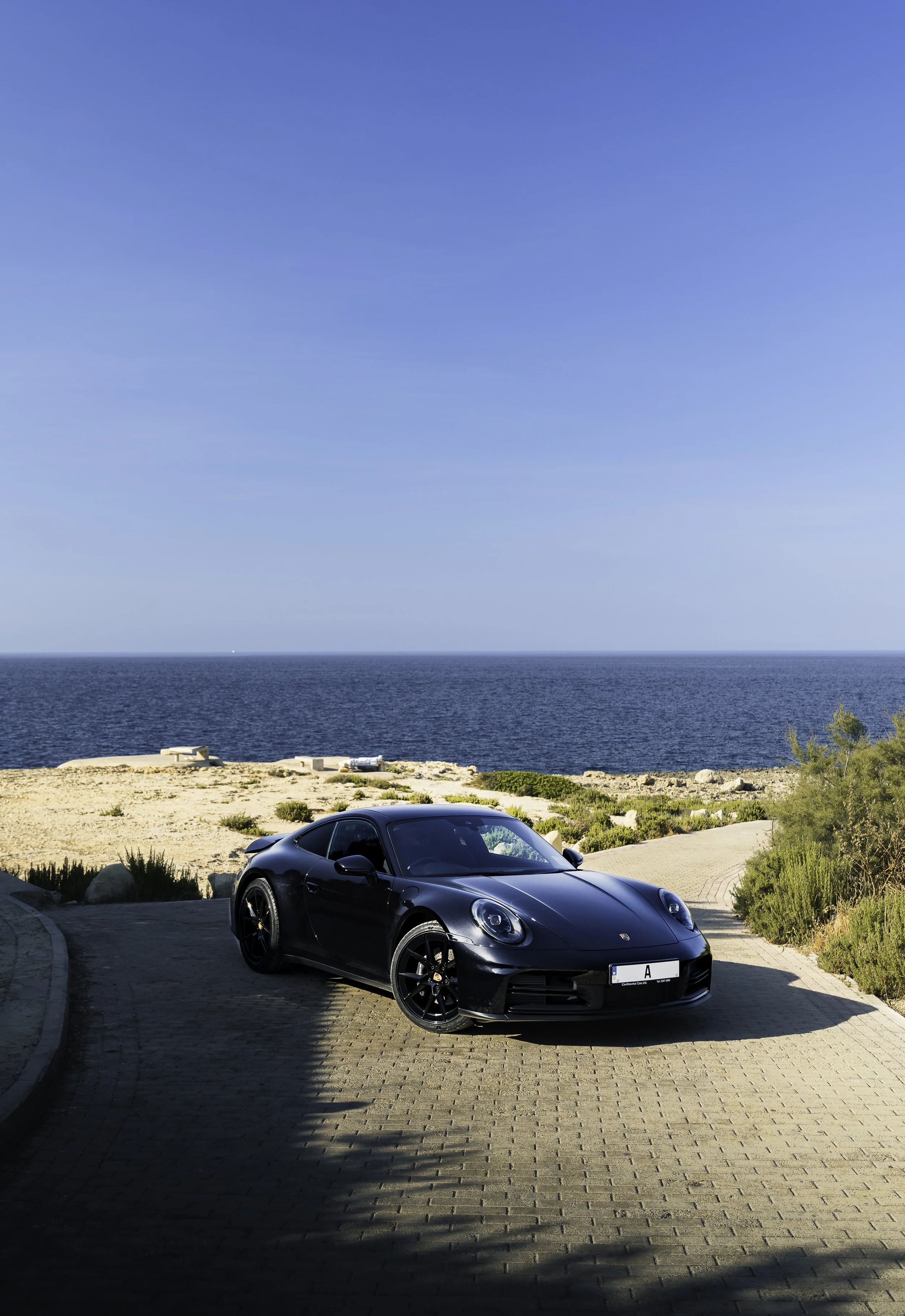 Black sports car parked on a cobblestone driveway near a cliff overlooking the ocean with clear blue sky overhead.