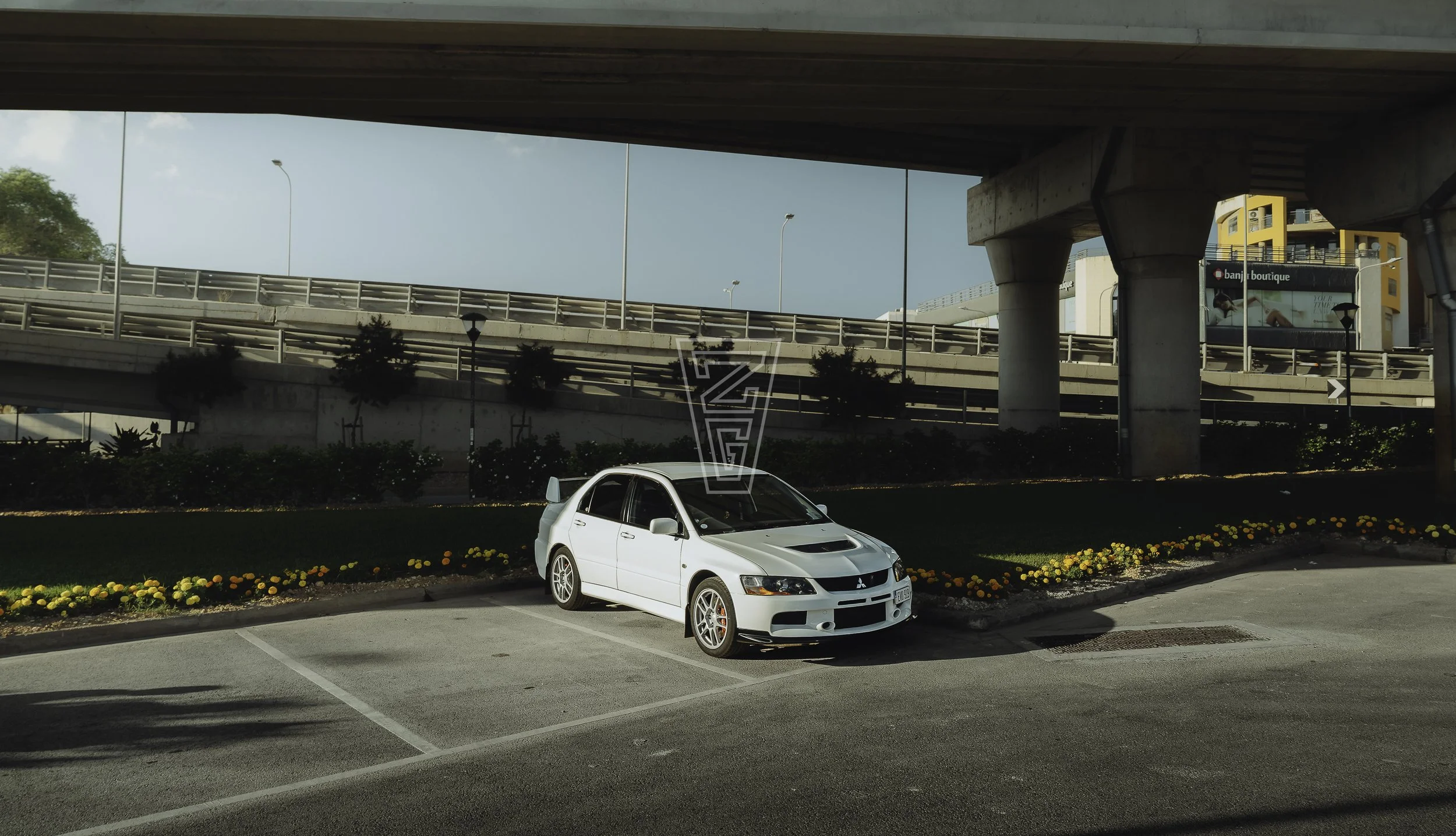 A white Mitsubishi Lancer Evolution car parked in a parking lot with a flower bed border, under an overpass with multiple levels of roads and a billboard sign in the background.
