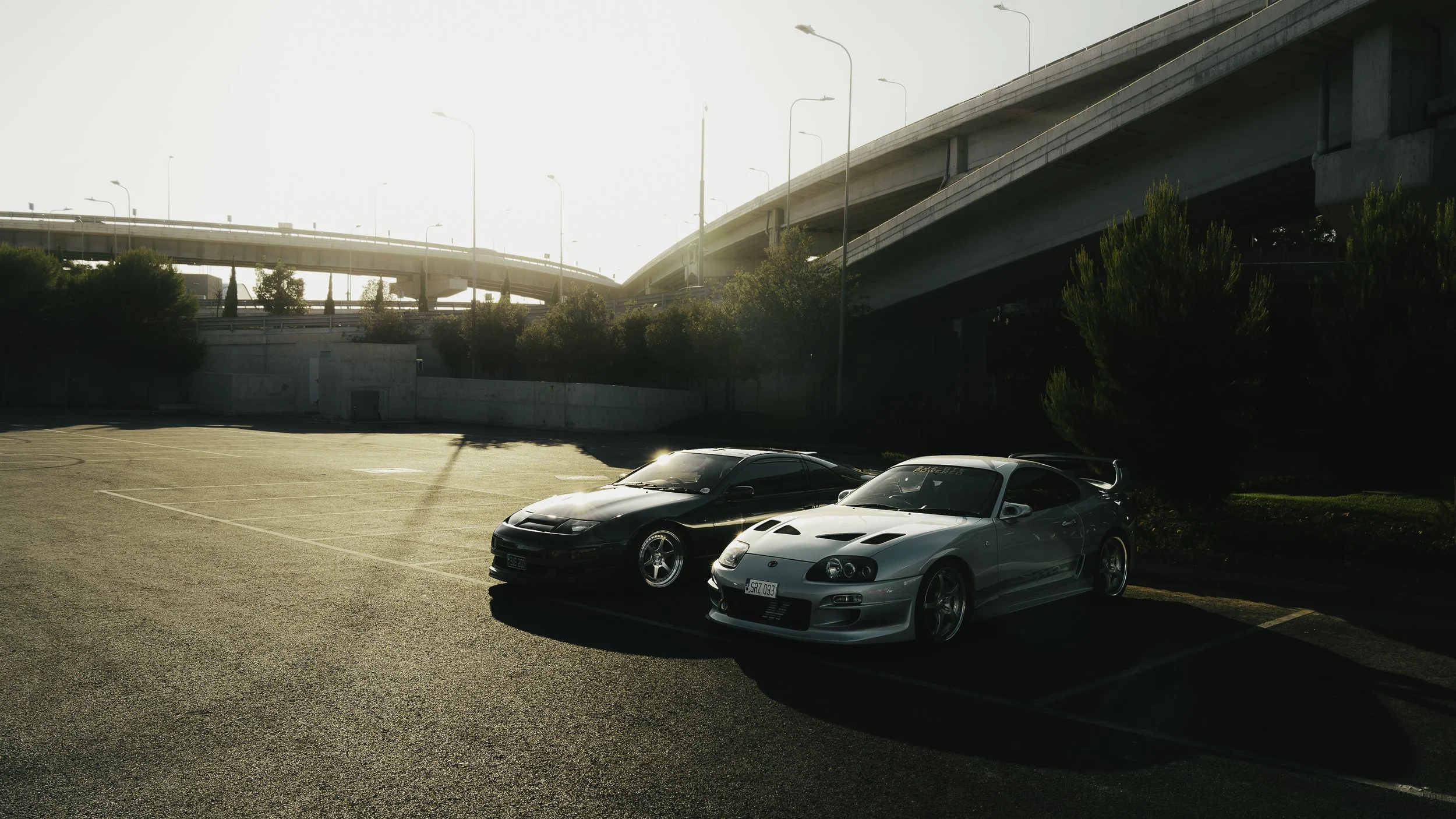 A parking lot with two sports cars, one black and one silver, parked underneath an overpass during sunset.