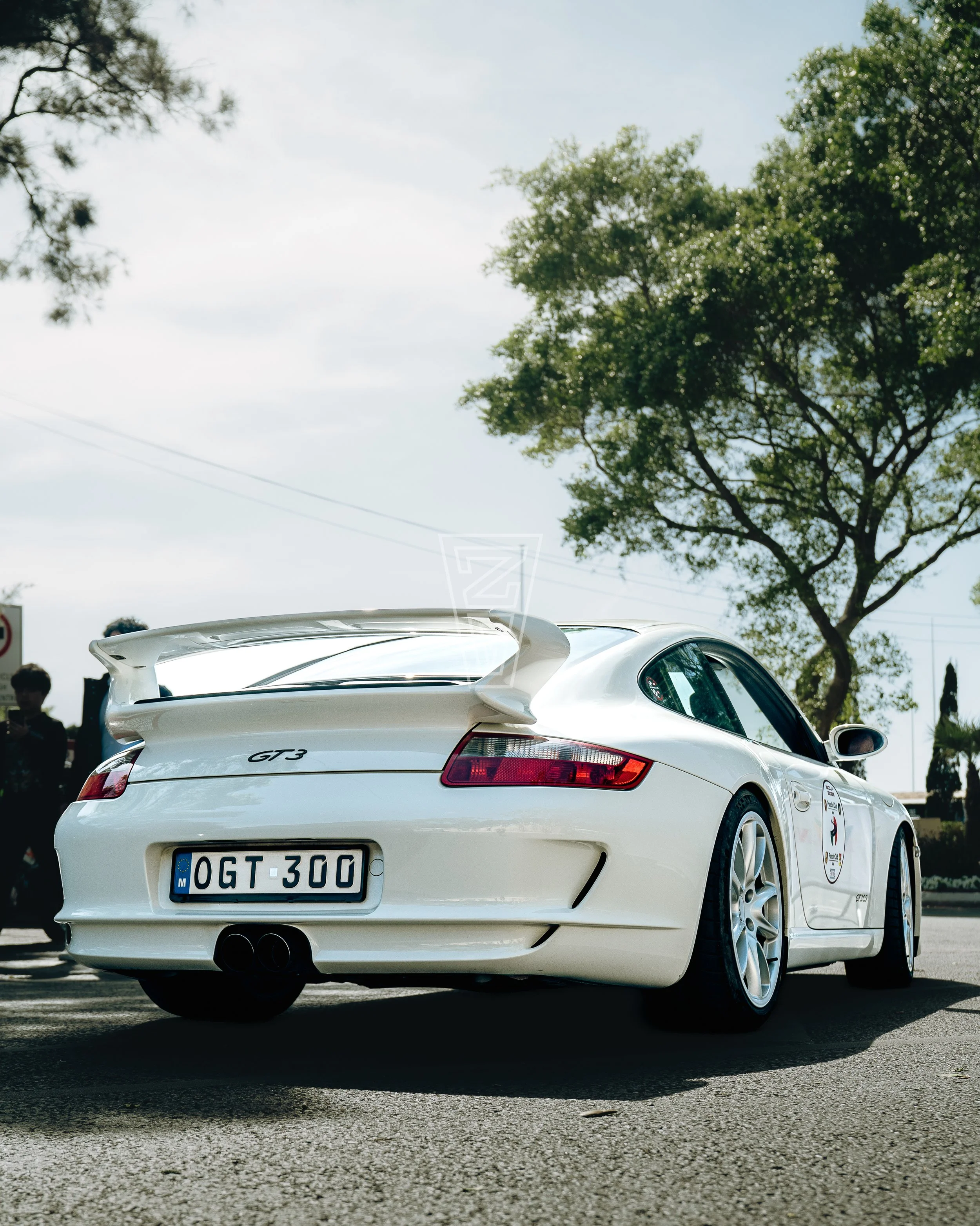 A white sports car with a large rear spoiler, parked on a paved surface with trees and people in the background, and a European license plate reading 'OGT 300'.