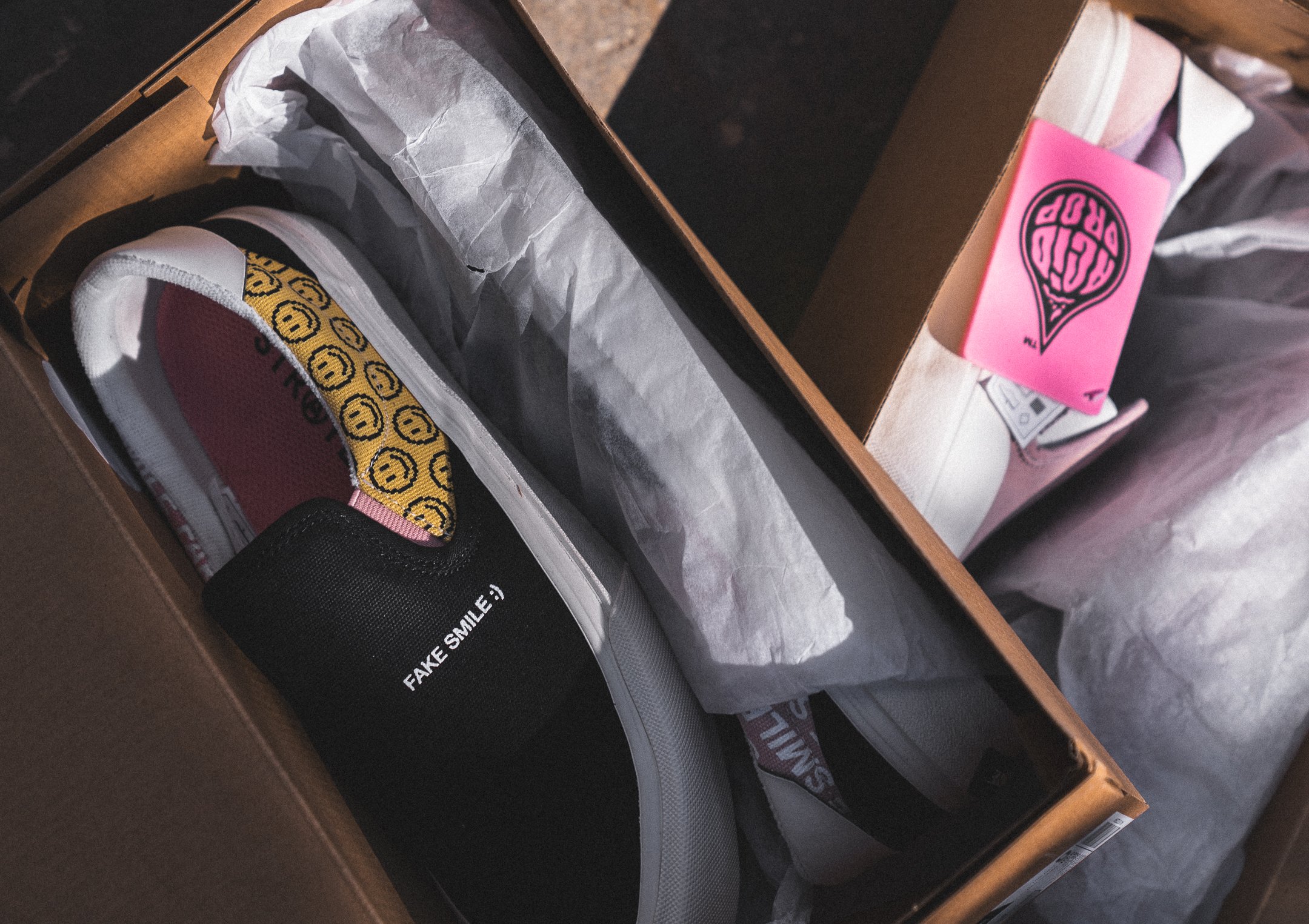 Black Straye slip-on sneakers with Fake Smile text and yellow smiley face patterns in a cardboard box.