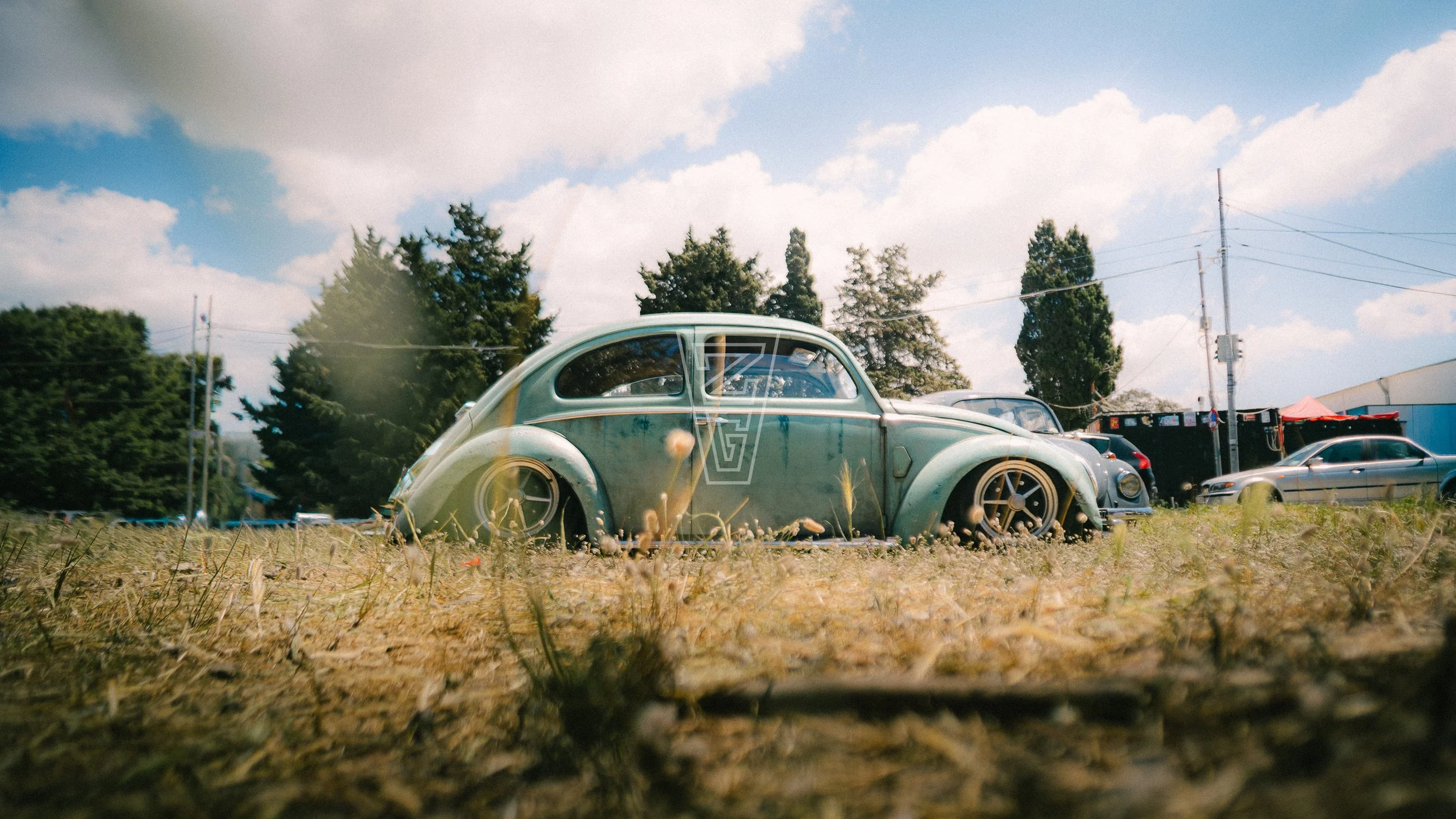 An old, weathered Volkswagen Beetle car parked on a grassy lot with other vehicles and trees in the background under a partly cloudy sky.