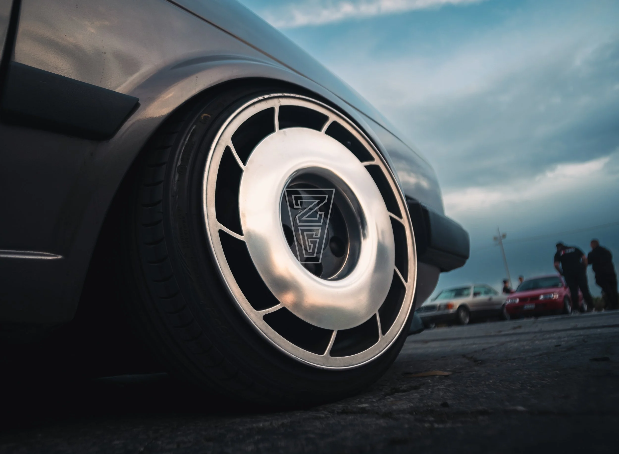 Close-up of a car wheel with a distinctive multi-spoke rim design, part of a silver or gray vehicle parked in a lot with other cars and people visible in the background under a cloudy sky.
