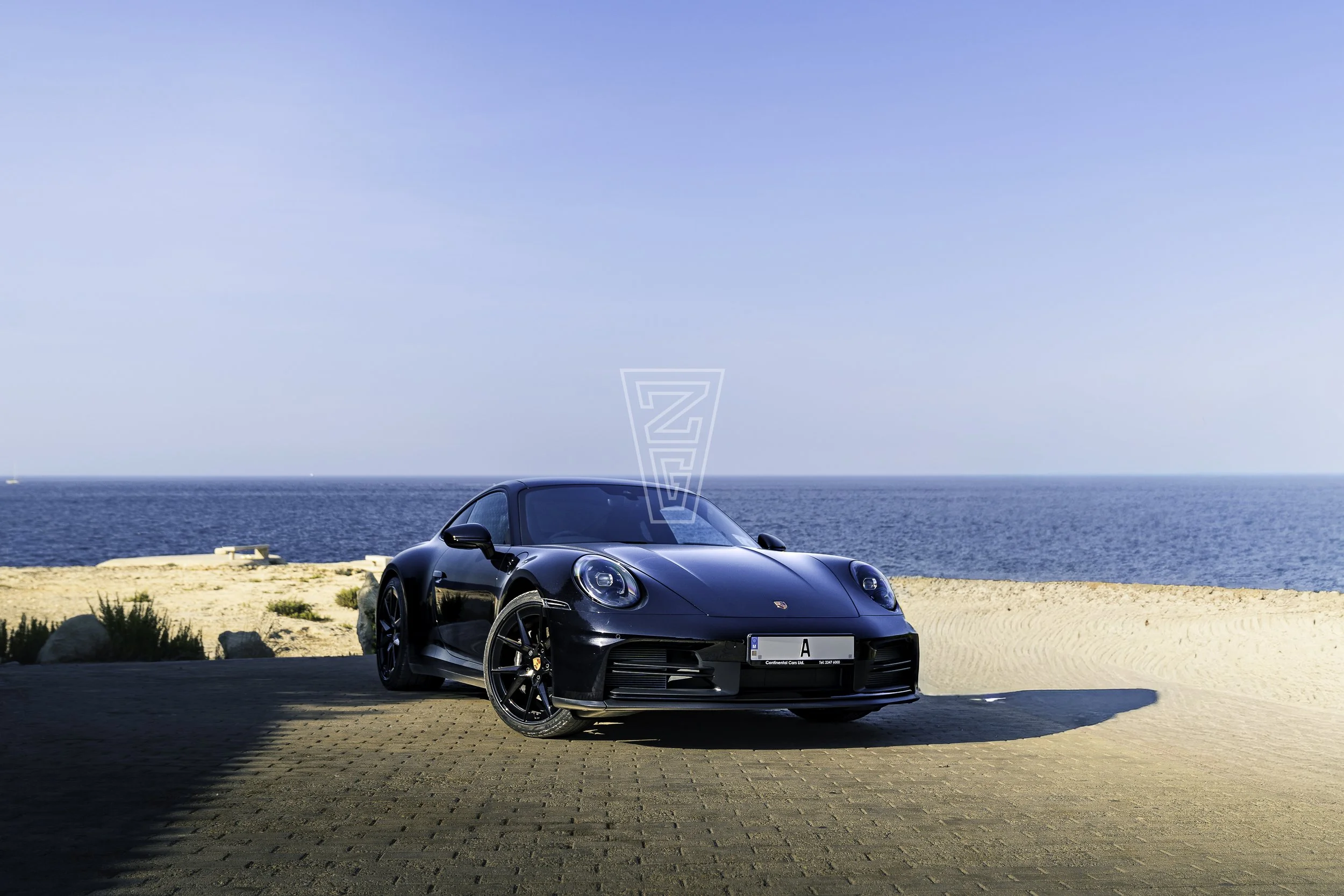 A black Porsche sports car parked on a paved area near the beach with the ocean and clear blue sky in the background.