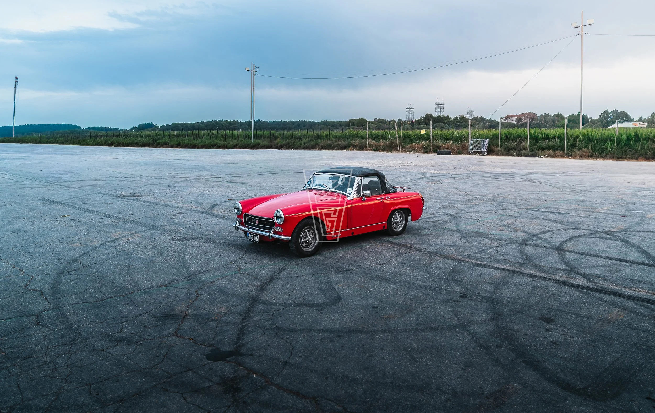 Red vintage convertible car parked on an empty cracked pavement in a rural area under a cloudy sky with utility poles and green fields in the background.