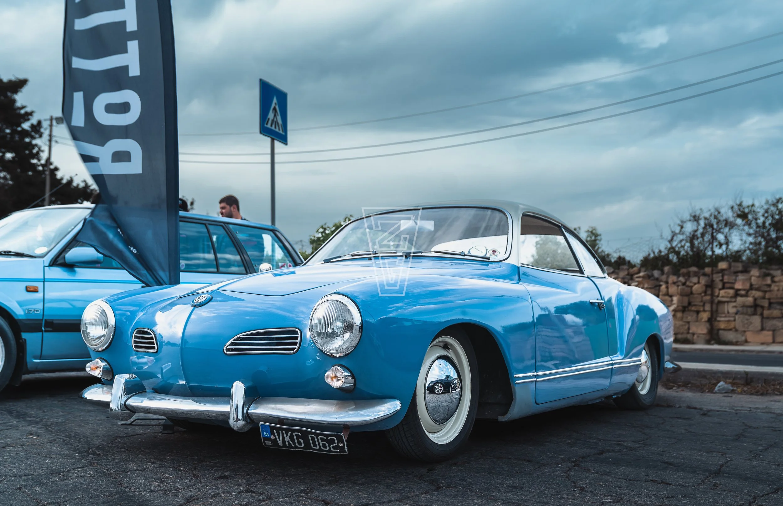 A vintage blue Volkswagen Karmann Ghia parked outdoors during daytime under a cloudy sky, with a man and other cars visible in the background.