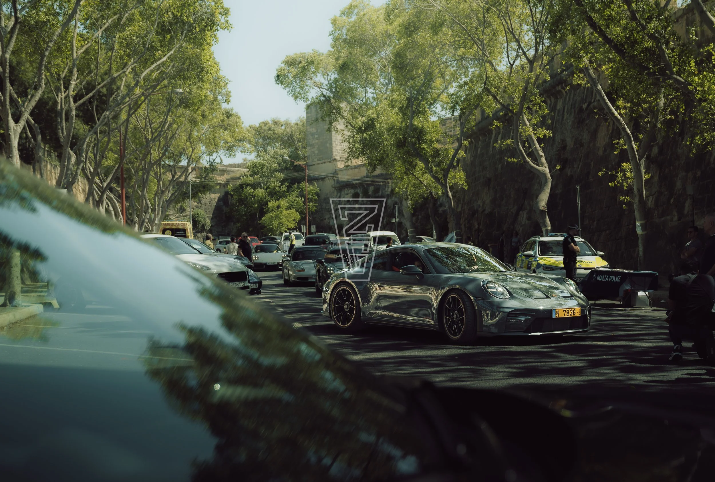 A parking lot with multiple parked cars, people walking, police and ambulance vehicles, and lush green trees, with old stone walls and a tall building in the background.