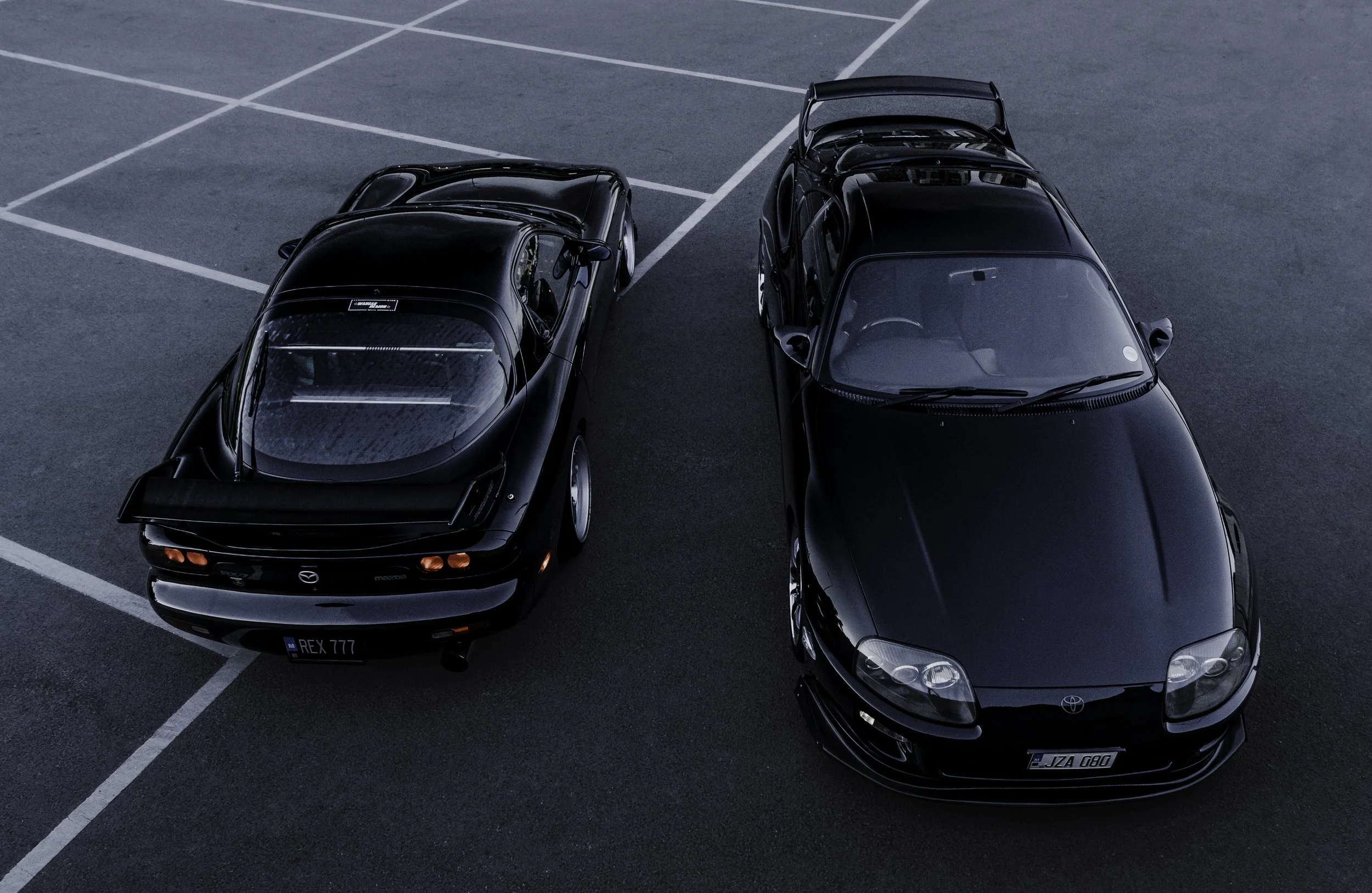 Top-down view of two black cars parked in a parking lot with white lines, one is a Mazda and the other a Toyota.