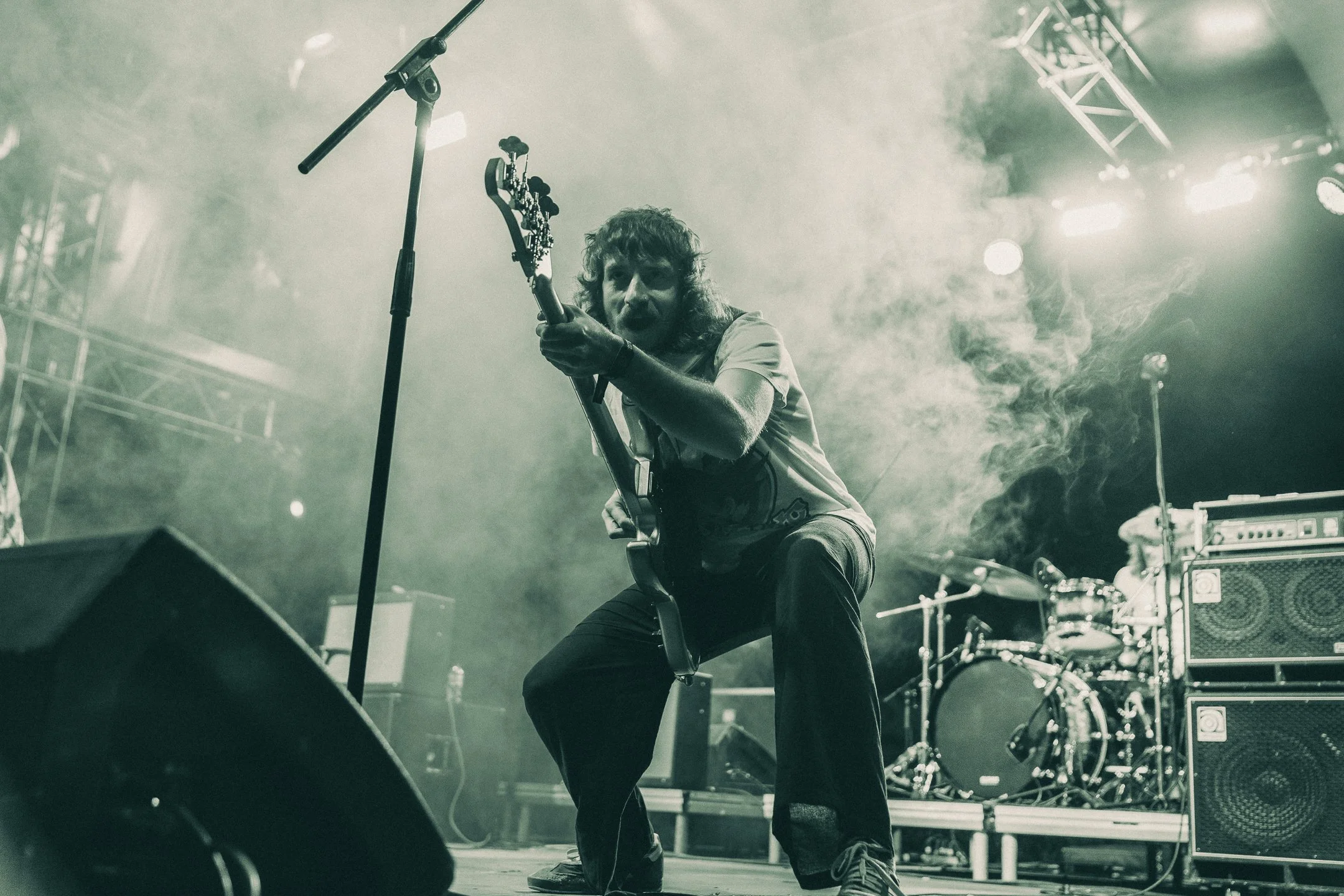 A musician performing on stage with a guitar, crouching down and looking directly at the camera amidst stage lights and smoke, with drums and amplifiers in the background.