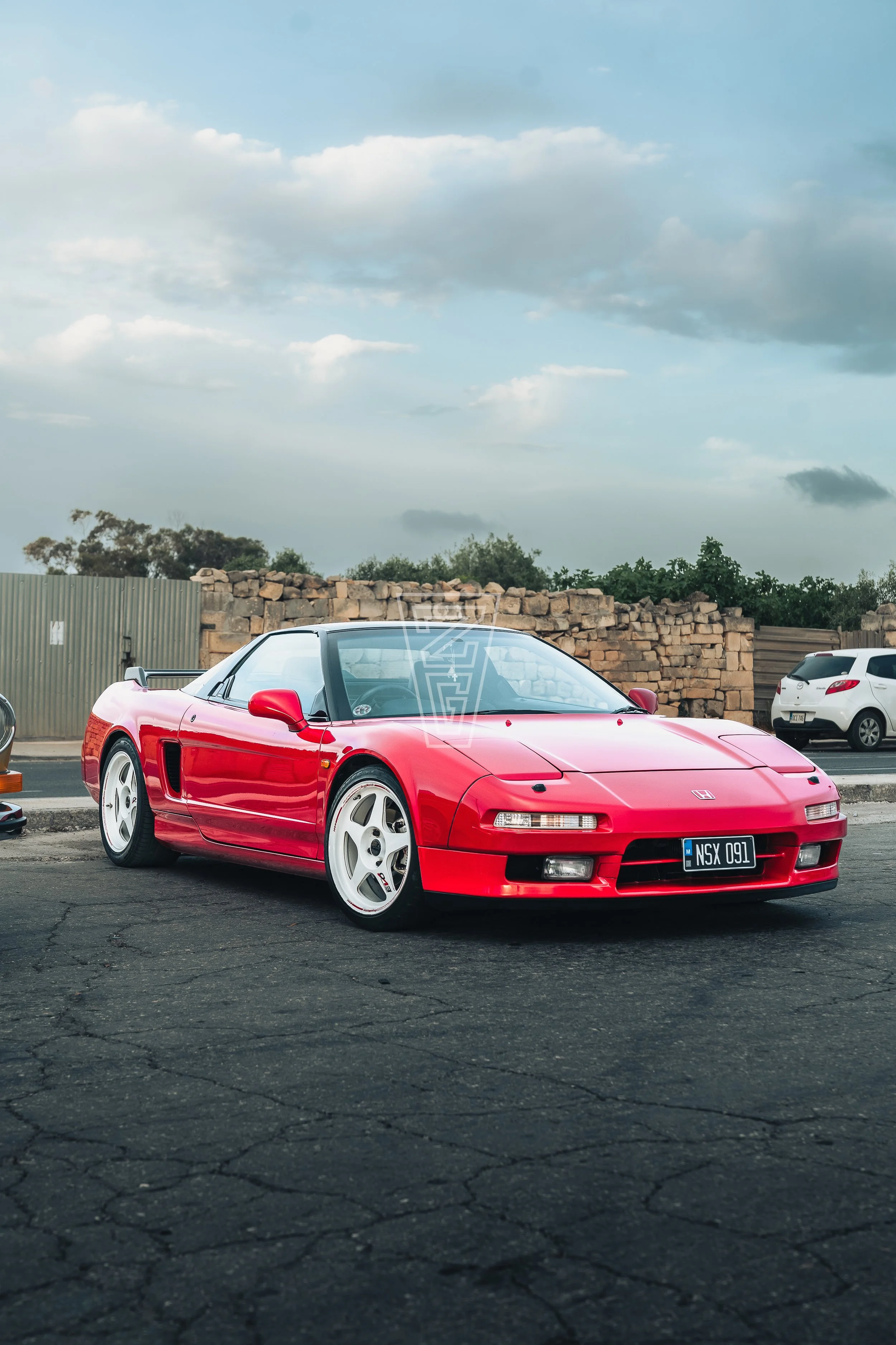 Red Honda NSX sports car parked on asphalt lot with other vehicles and brick wall in background, overcast sky.