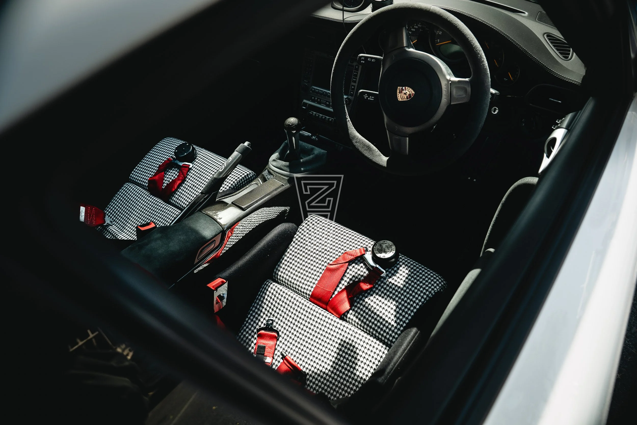 The interior of a sports car, showing a steering wheel with a Porsche emblem, racing seats with checkered patterns, harnesses, and various dashboard controls.