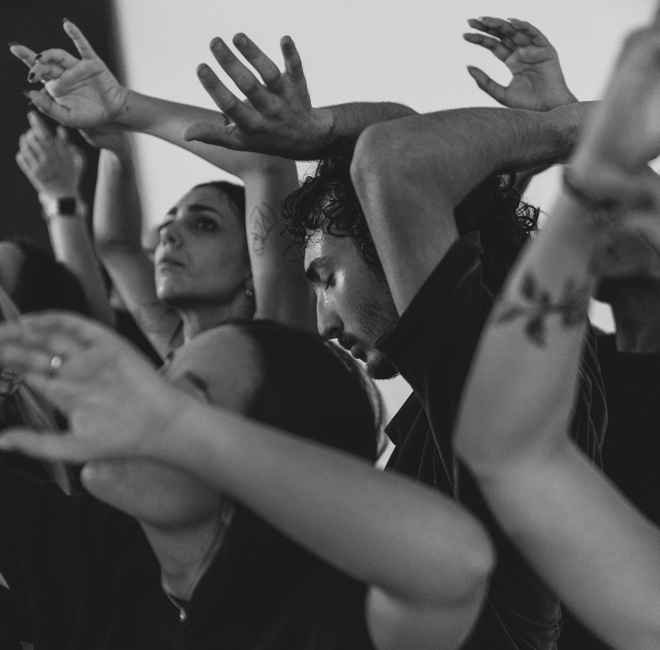 Black and white photo of diverse group of people with their hands raised, some with eyes closed or looking upward, suggesting a moment of contemplation or participation in an activity.