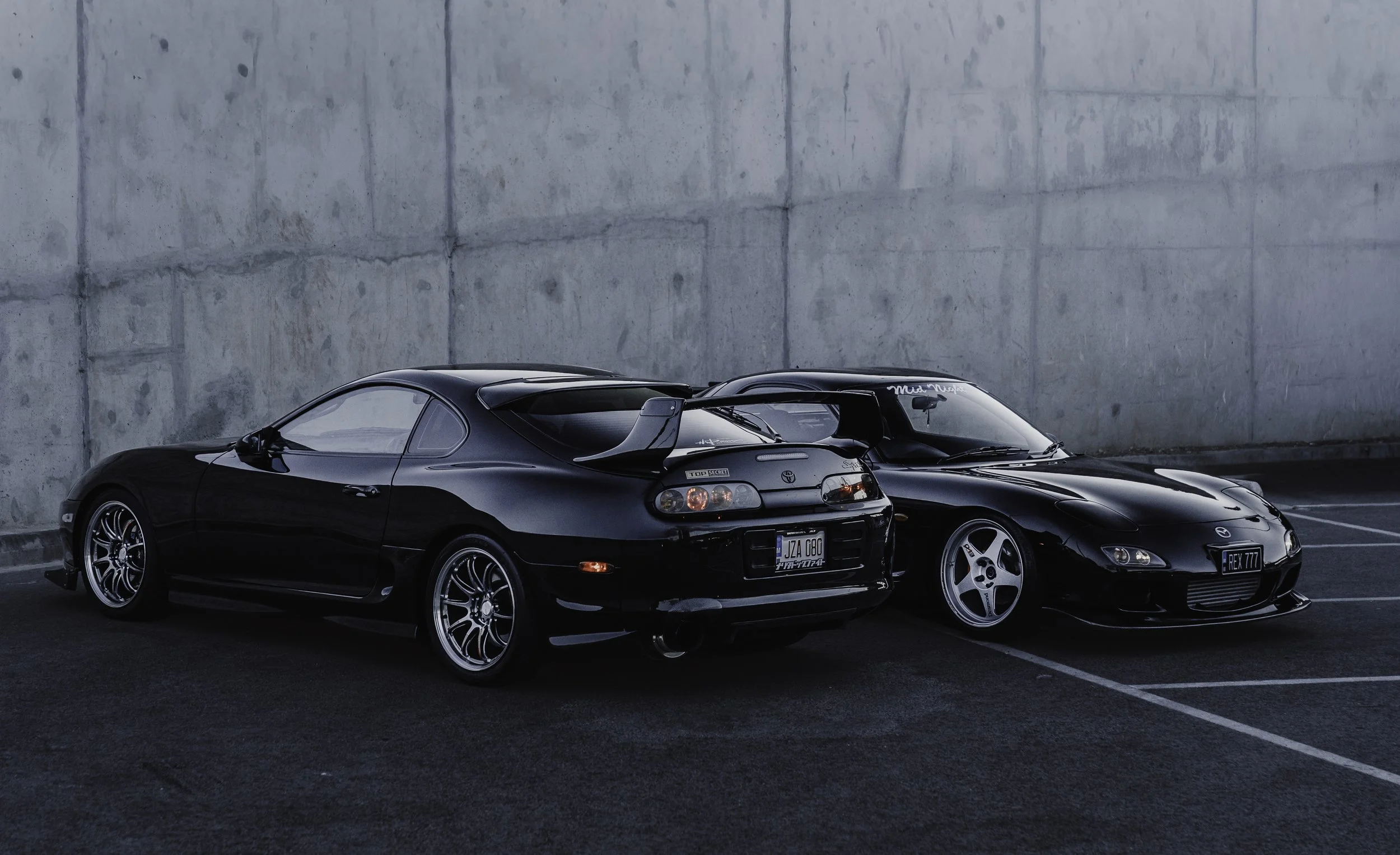 Two black sports cars parked against a concrete wall at night.