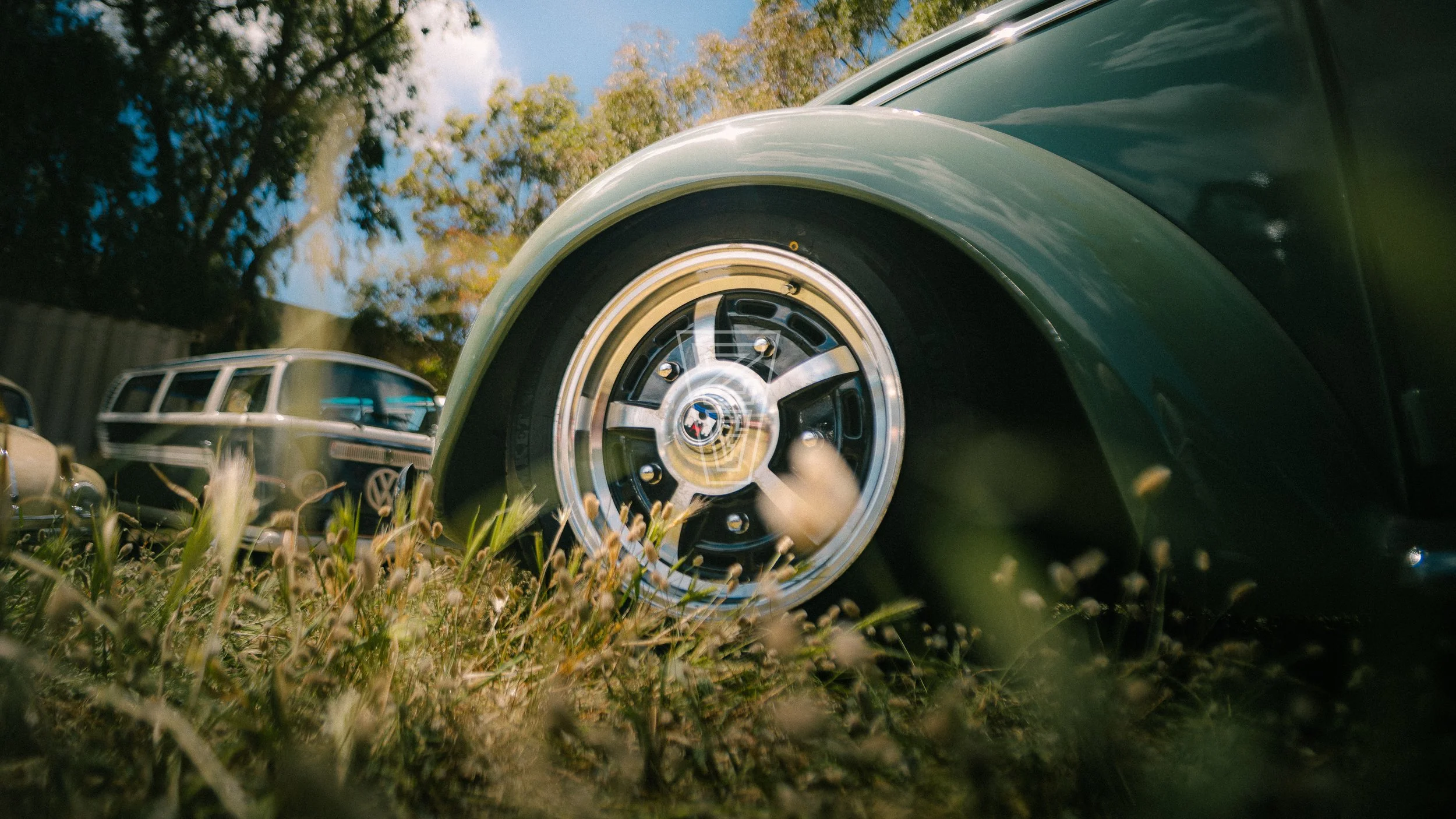 Close-up of the front wheel of a vintage green car parked on grass with other classic cars in the background and trees under a partly cloudy sky.